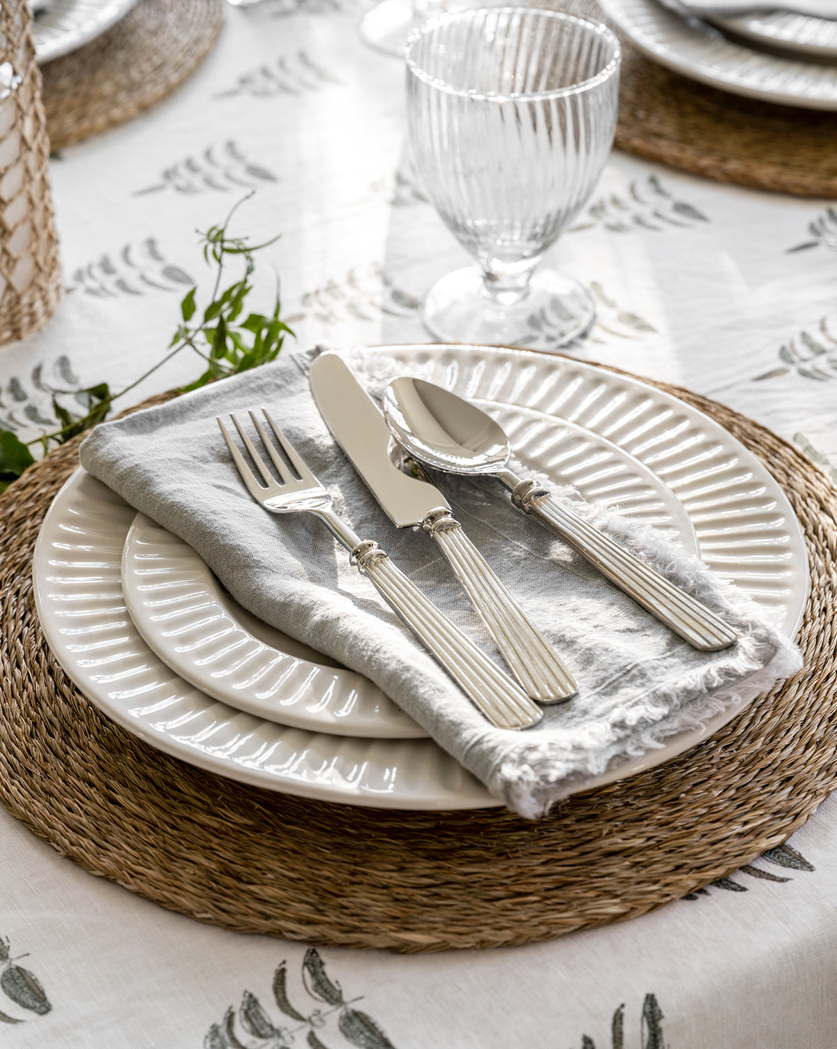 A close-up of a table setting with two stacked white plates, a gray napkin, the McNully Flatware (Set of 5) with textured handles, a clear glass, and a woven placemat on a white green-leaf-patterned tablecloth by McGee & Co.