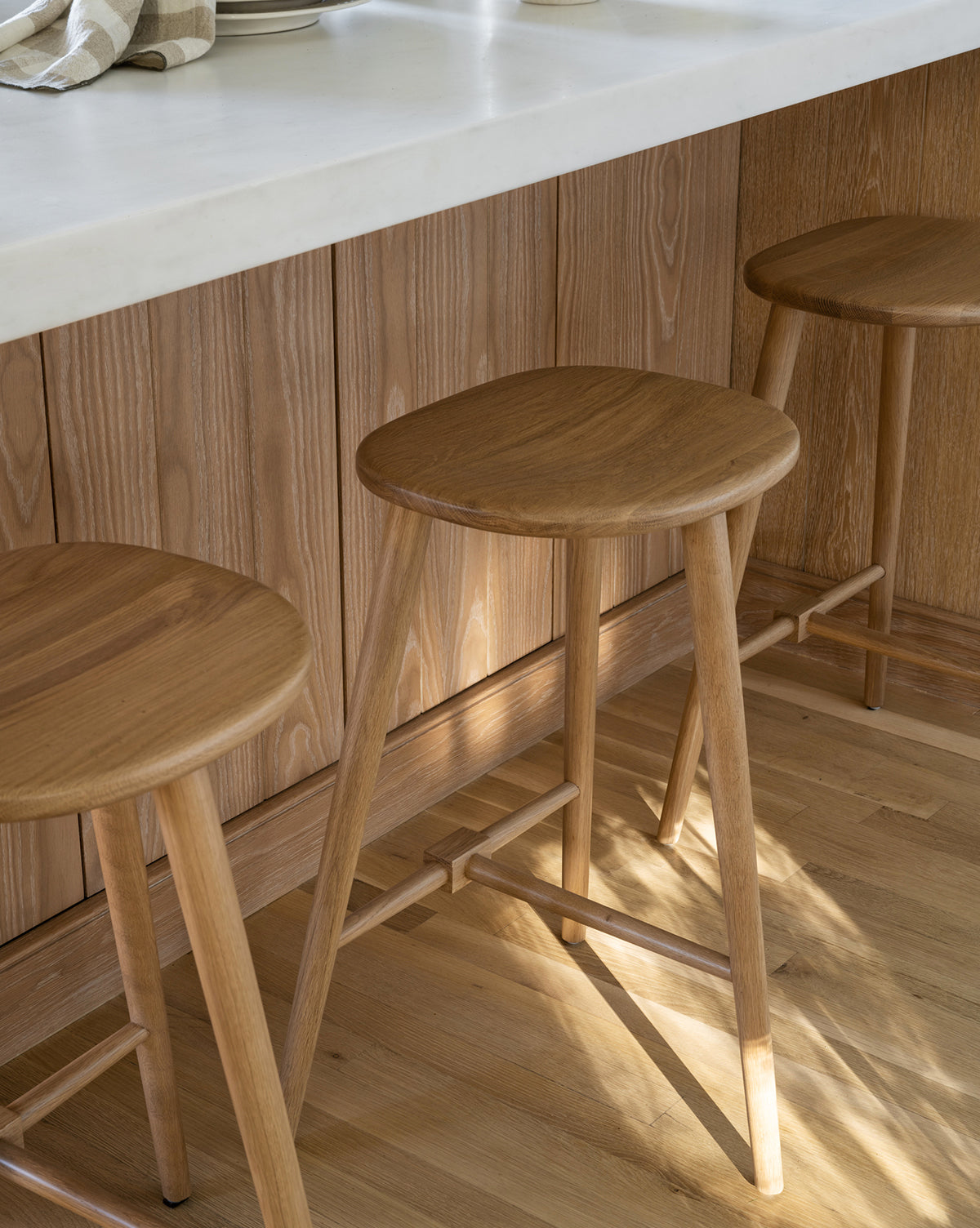 Three Fielder Counter Stools with round wooden seats and simple legs sit at a light oak kitchen counter topped with white, by McGee & Co. Sunlight streams in, casting gentle shadows across the wooden floor.