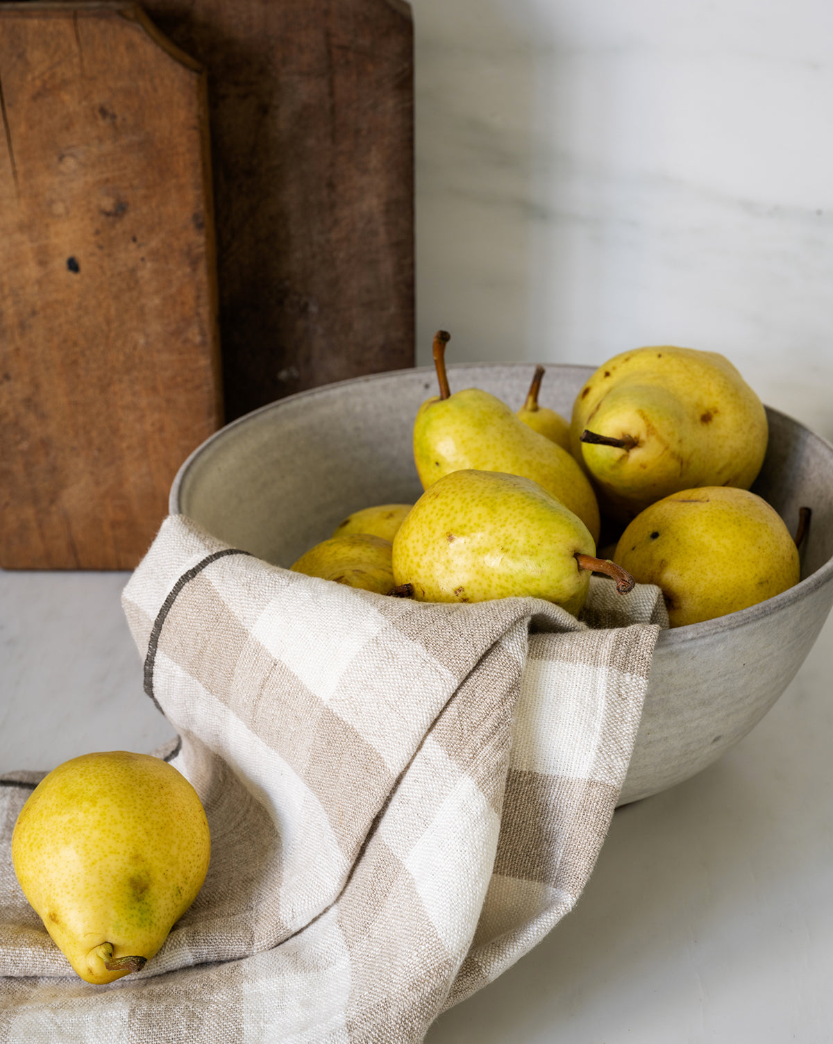 A bowl of yellow pears sits on a countertop, partially covered with the Charlie Check Tea Towel. One pear rests outside the bowl, while wooden boards in the background create a cozy table setting, by McGee & Co.