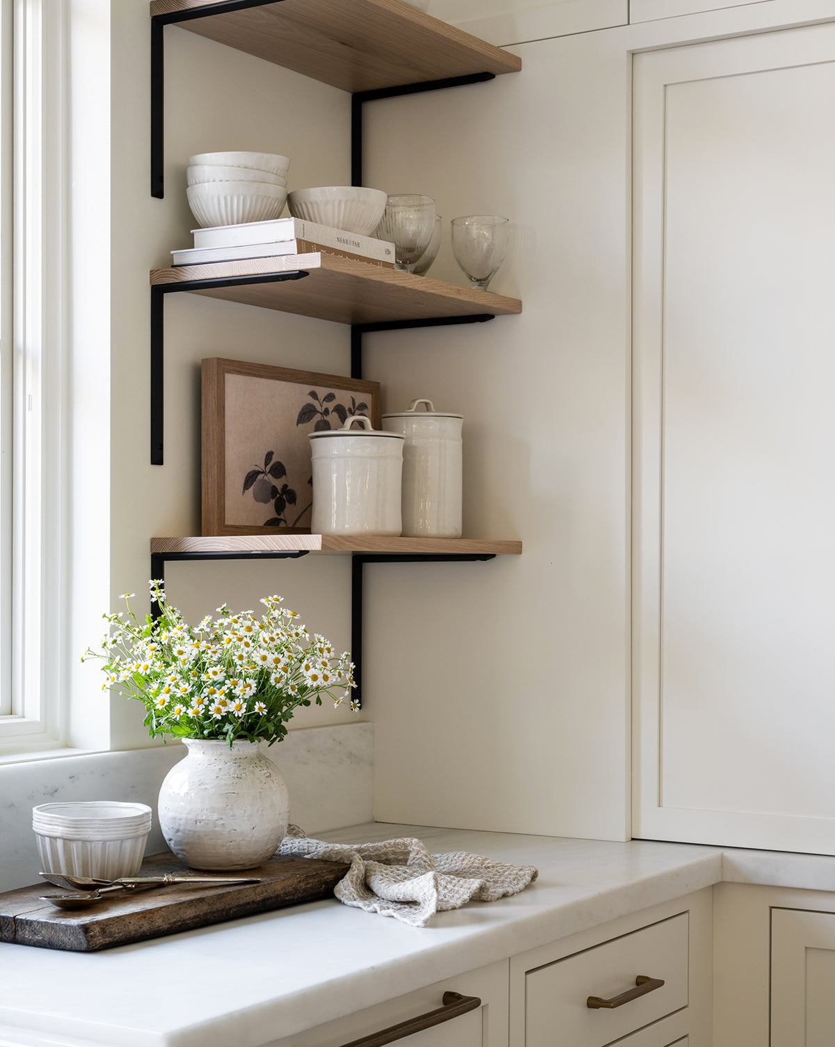 A bright kitchen corner with floating wooden shelves displays white dishes and glassware. On the marble countertop sit a vase of daisies, a wooden tray, and a Handled Ceramic Canister next to a folded dish towel.
