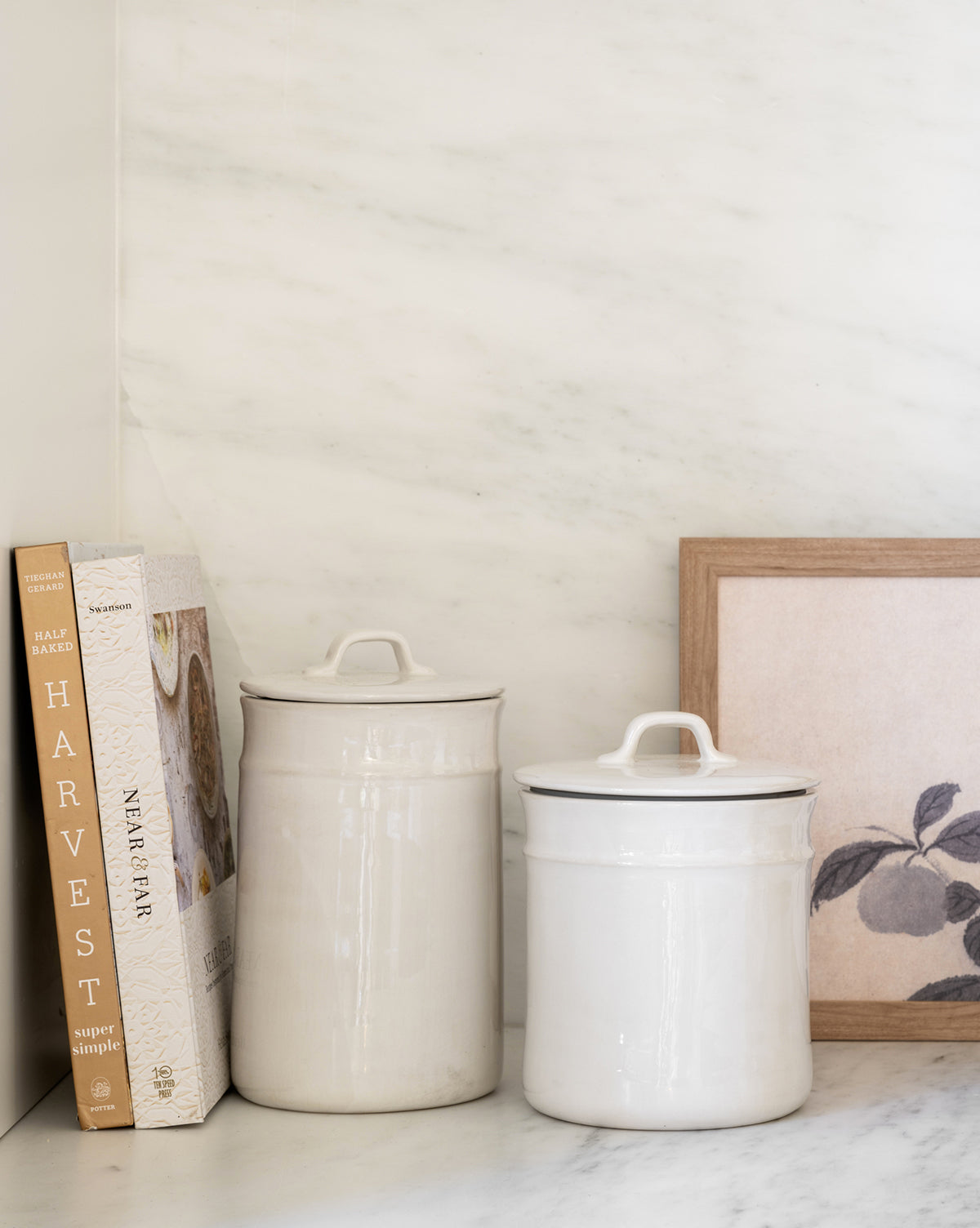 Two Handled Ceramic Canisters sit on a marble countertop beside two cookbooks and a framed fruit art print, adding stylish storage and modern flair to the kitchen against a white marble wall.