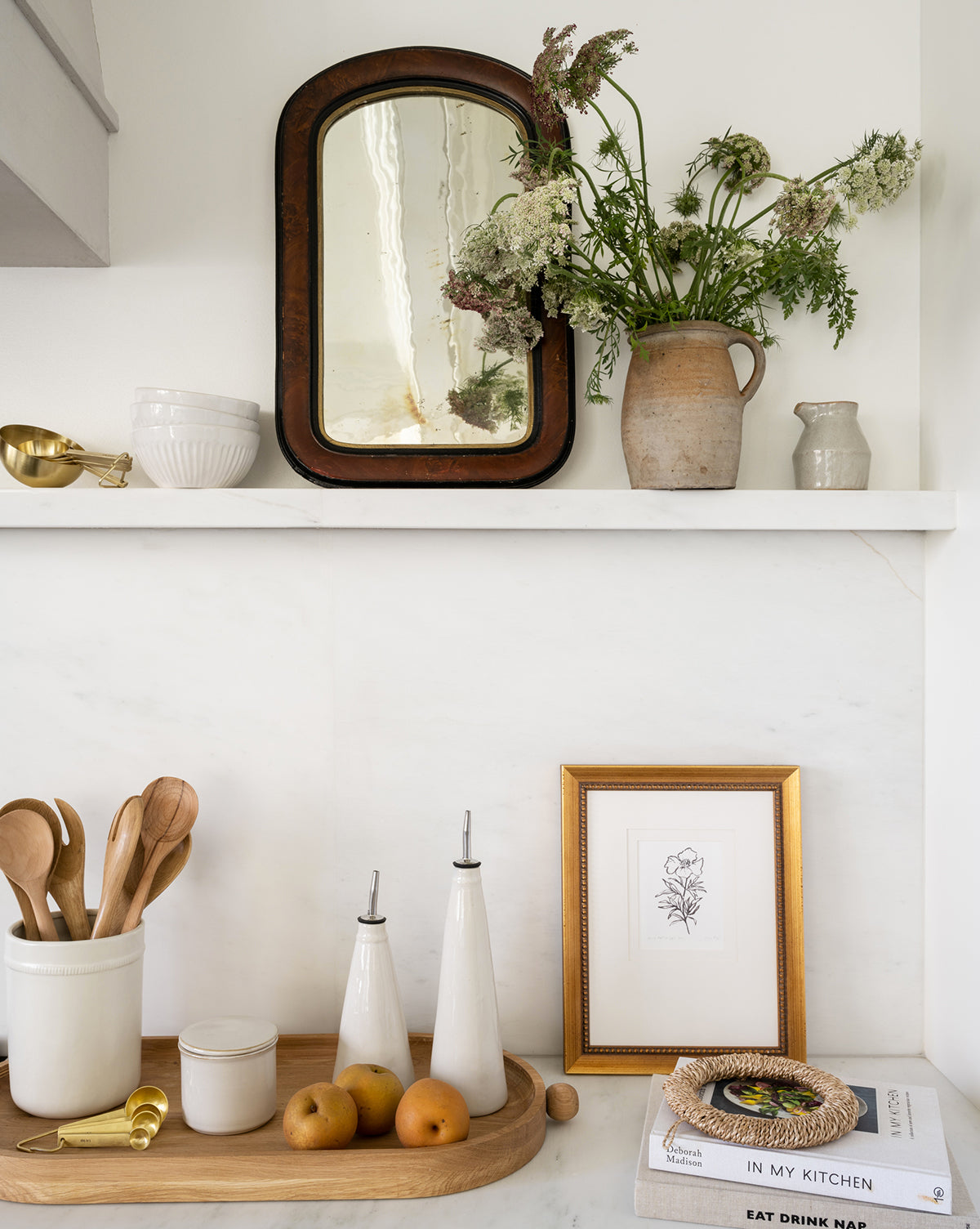 A kitchen shelf styled with a rustic mirror, wildflowers in a white vase, the Dashed Ceramic Crock holding wooden utensils, pears, olive oil bottles, a framed line drawing, and stacked cookbooks on a light countertop—perfect for kitchen decor.