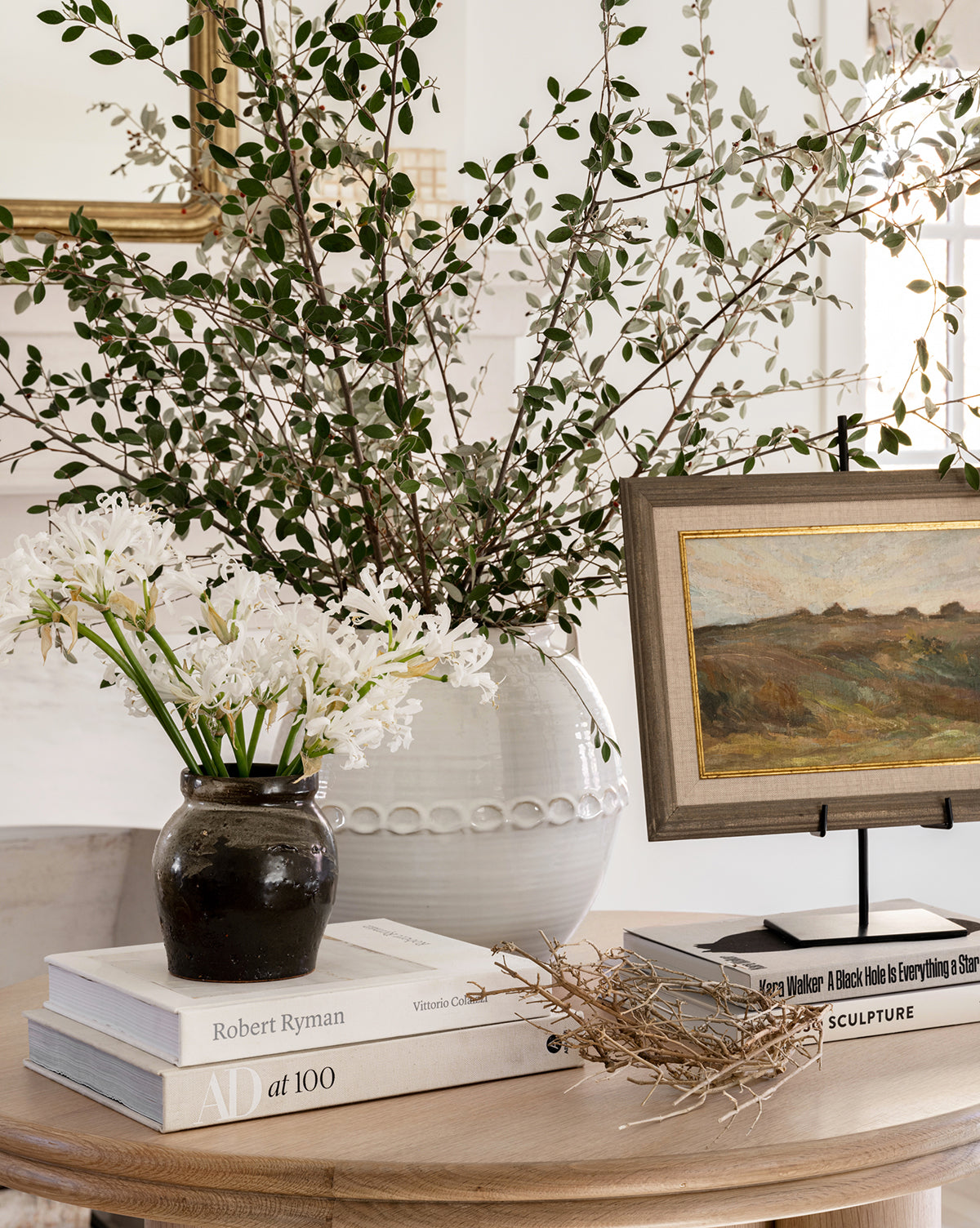 A round wooden table holds stacked art books, the Dark Glazed Ceramic Vase with white flowers, a larger white vase with leafy branches, a dried branch, and a framed landscape painting on a stand.
