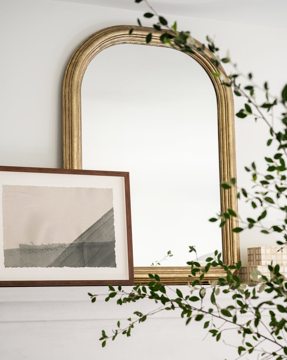 A large arched Giada Wall Mirror with an elegant moulded frame is mounted on a white wall above a shelf, with a framed abstract artwork, a small decorative object, and green leafy branches arranged in front, by McGee & Co.