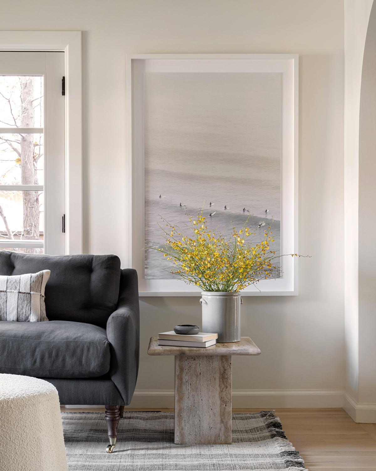 A cozy living room with a dark gray sofa, a small wooden side table with yellow flowers, and a large framed museum-grade original photograph called Surf Rider displayed on a light-colored wall.
