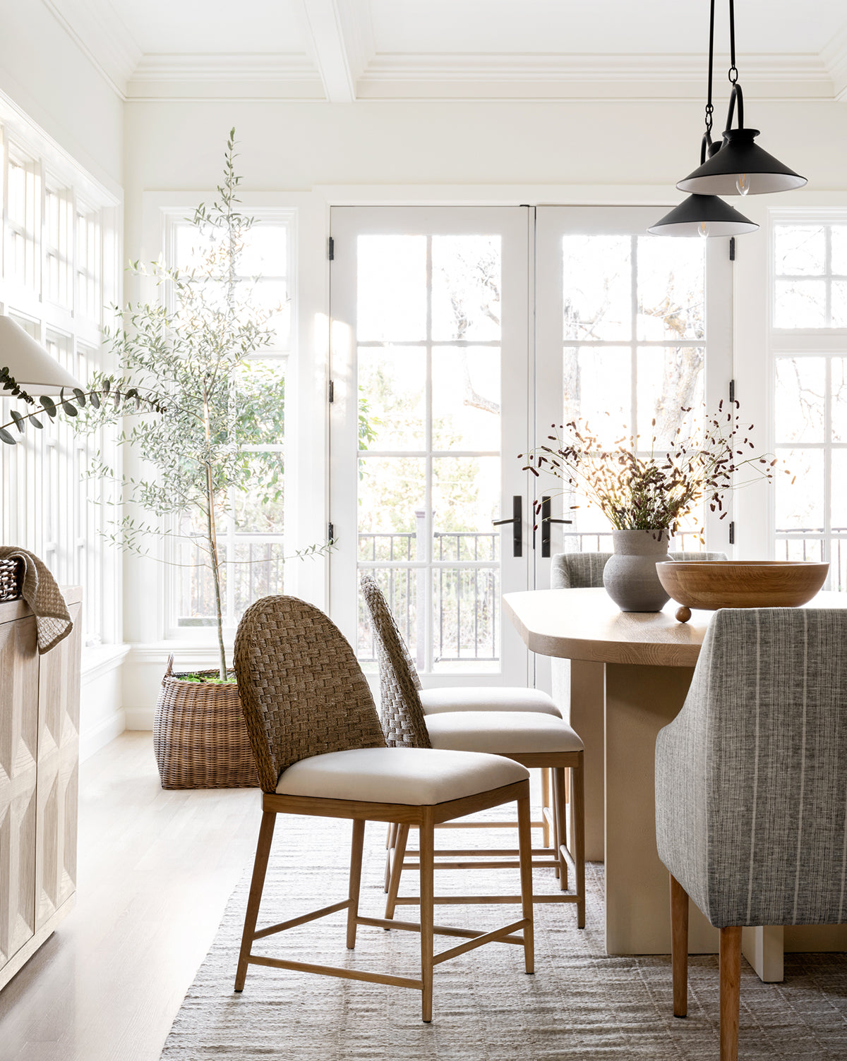 A bright dining room with large windows and a solid oak table features the Molly Dining Chair, combining woven and upholstered seats, atop a patterned rug. Potted plants and decorative bowls add cozy, modern style bathed in natural light by McGee & Co.