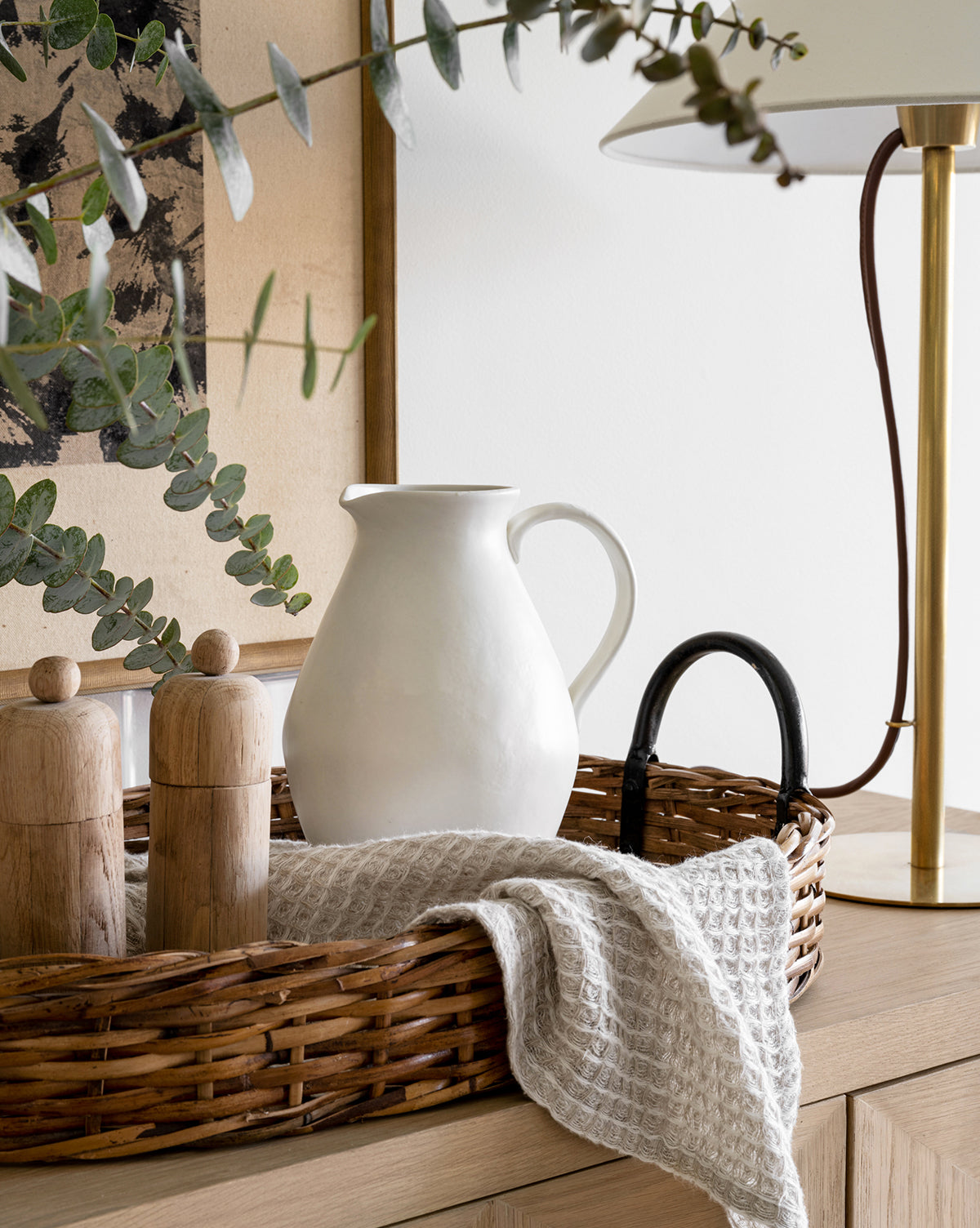 A wicker tray on wood holds a White Ceramic Pitcher, two wooden grinders, and a textured cloth; eucalyptus branches and a brass lamp sit nearby, by McGee & Co.