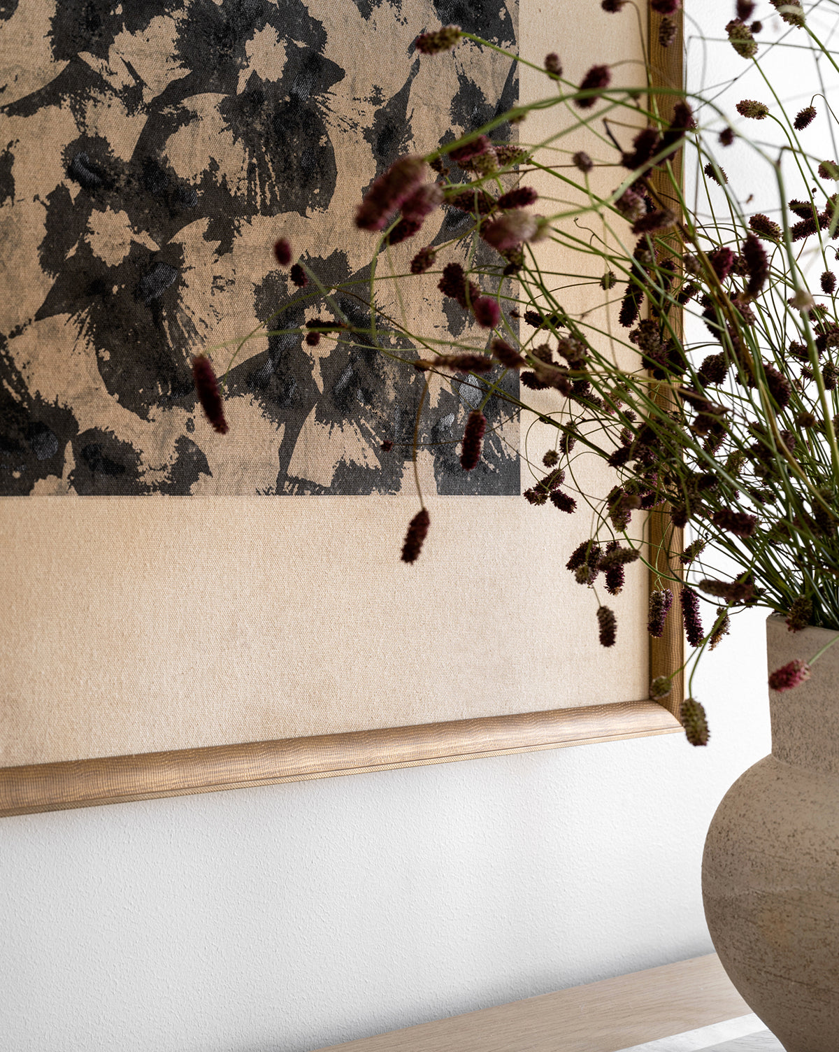 A close-up of the Woodblocked Botanical artwork in a gold frame hangs on the wall. In front, a stone-textured vase displays delicate stems with small, dark reddish flower clusters, creating a striking modern scene.