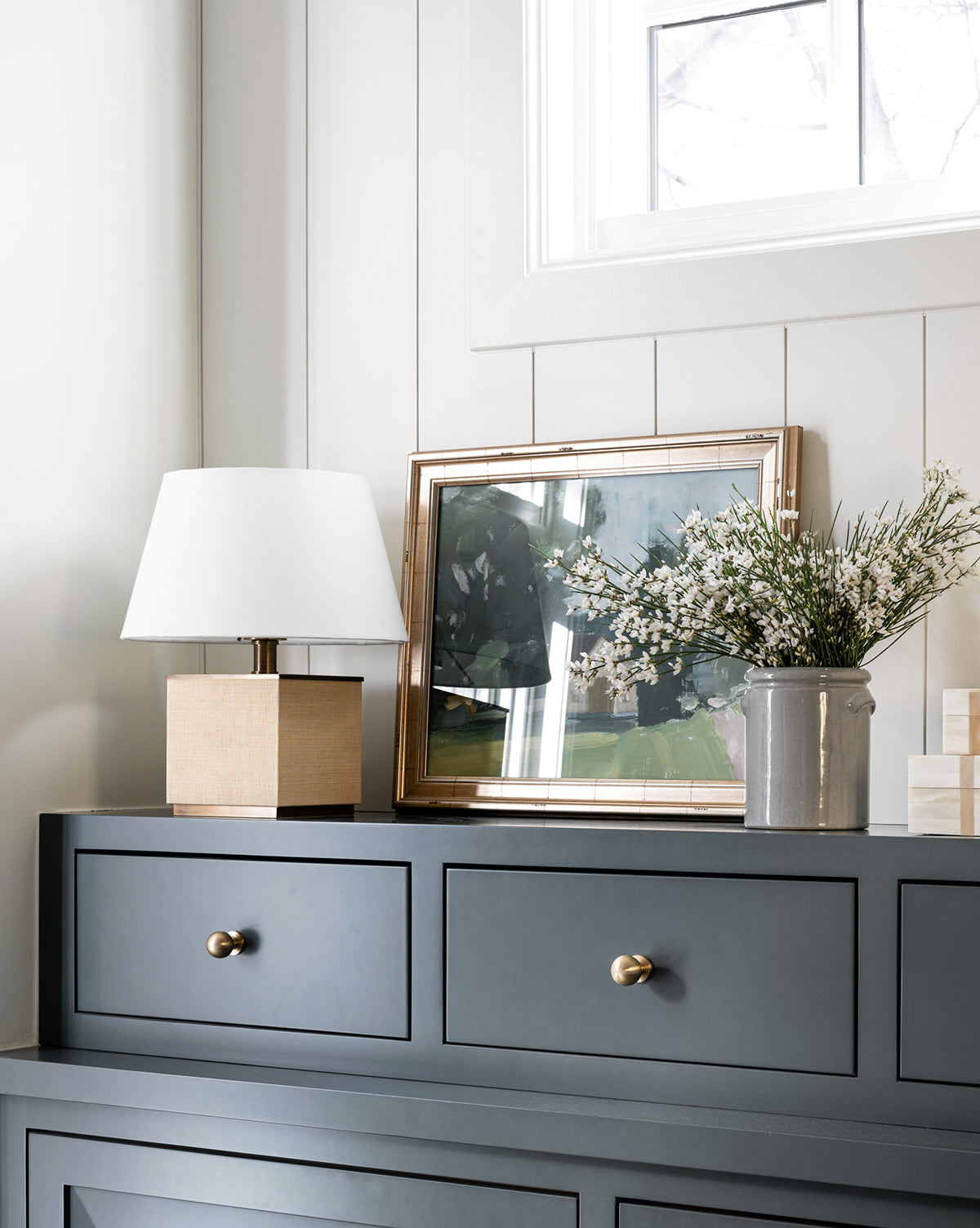 A dark blue dresser with gold knobs holds a square lamp, a gray vase of white flowers, and Flaxen Pasture, an abstract country painting. A window above provides natural light.