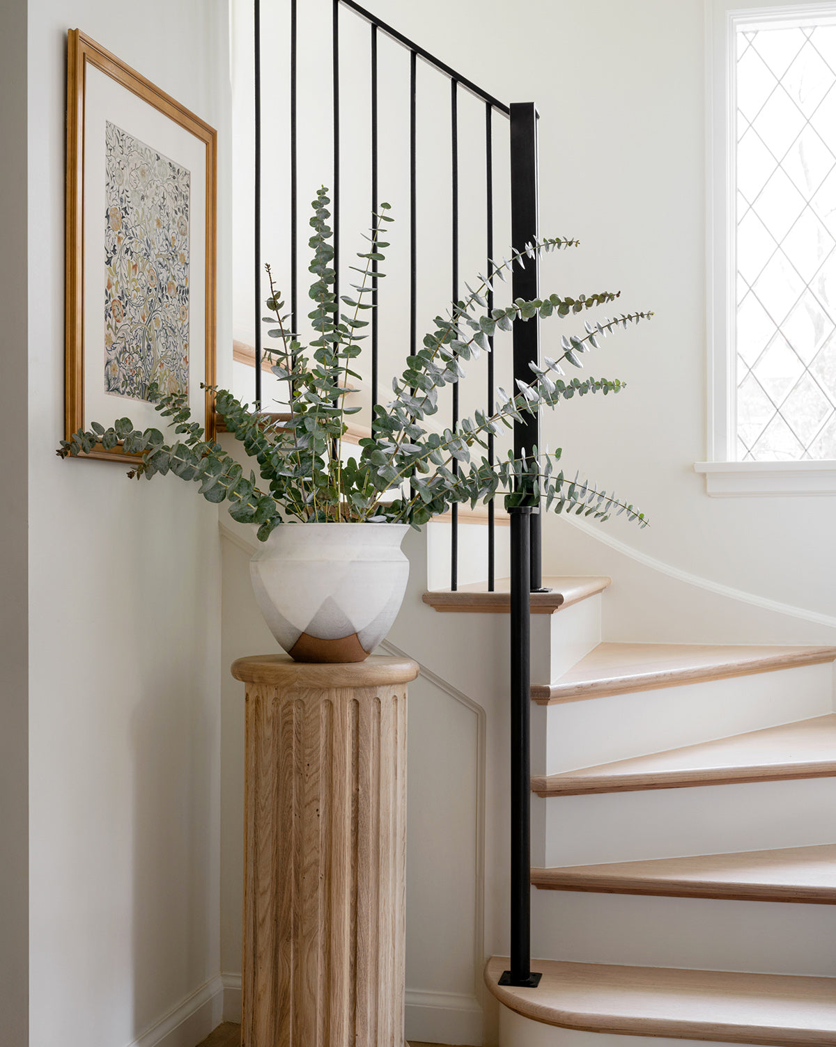 The Acanthus Portière, a large potted eucalyptus plant, sits on a wooden pedestal next to a curved staircase with light wood steps and black railing, in a bright room featuring white walls and antique gold-framed botanical art.
