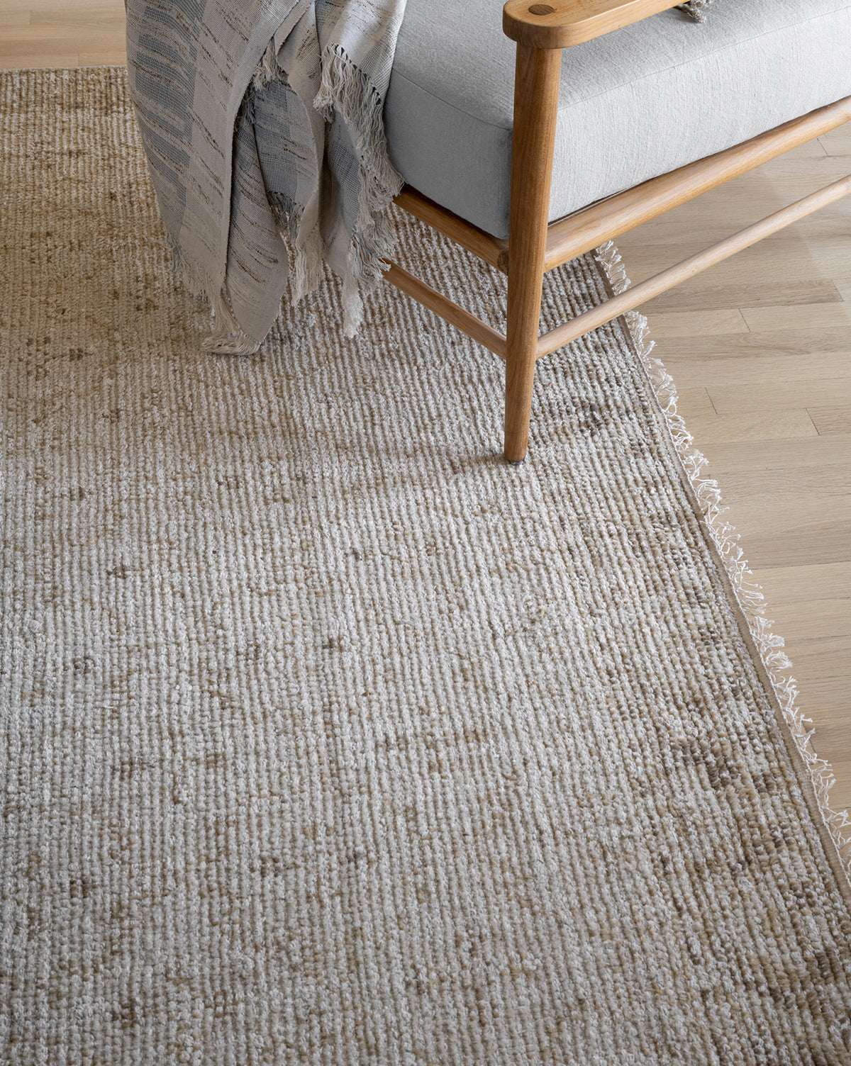 A close-up of the Mali Hand-Knotted Rug in beige on a light wood floor, beside a wooden chair with a grey cushion and a light, textured throw blanket draped over the arm, by McGee & Co.