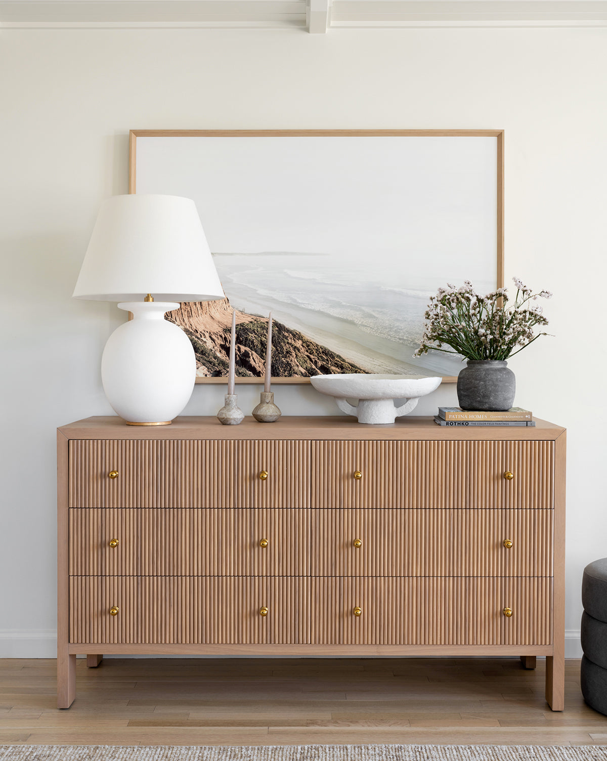 A wooden dresser with gold knobs holds a large white lamp, two candlesticks, a bowl, a gray vase with flowers, and the Distant Coastline framed wall art leaning against a white wall.