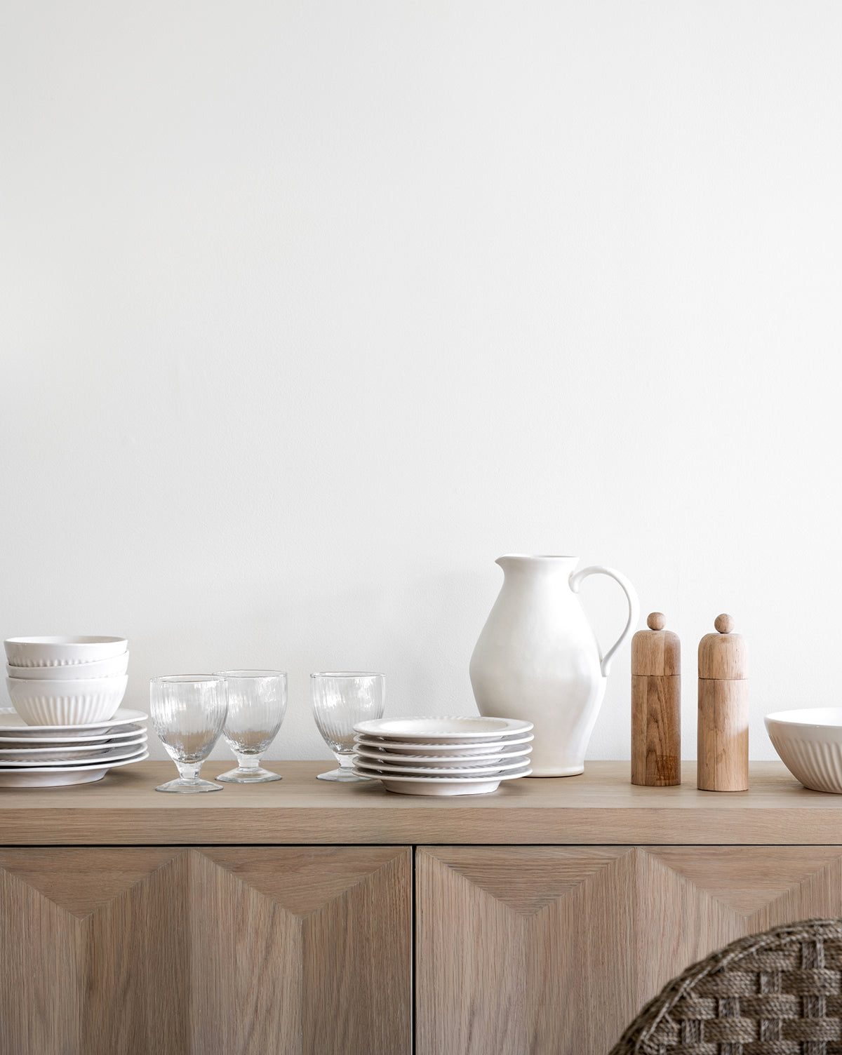 A minimalist wooden sideboard with fluted detailing displays stacked white dishes, clear glasses, a white ceramic pitcher, the Adele Bowl, and wooden salt and pepper grinders against a plain white wall, by McGee & Co.