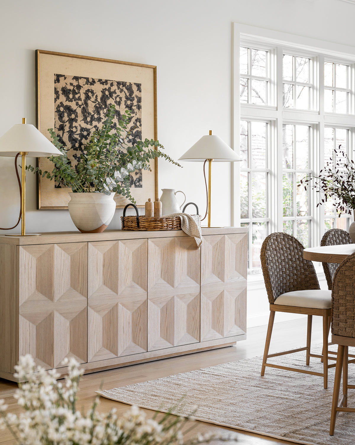A bright dining room features the Woodblocked Botanical giclee print in a gold frame, a textured wood sideboard, decorative vases, two modern lamps, wicker chairs around a round table, and large windows letting in natural light.