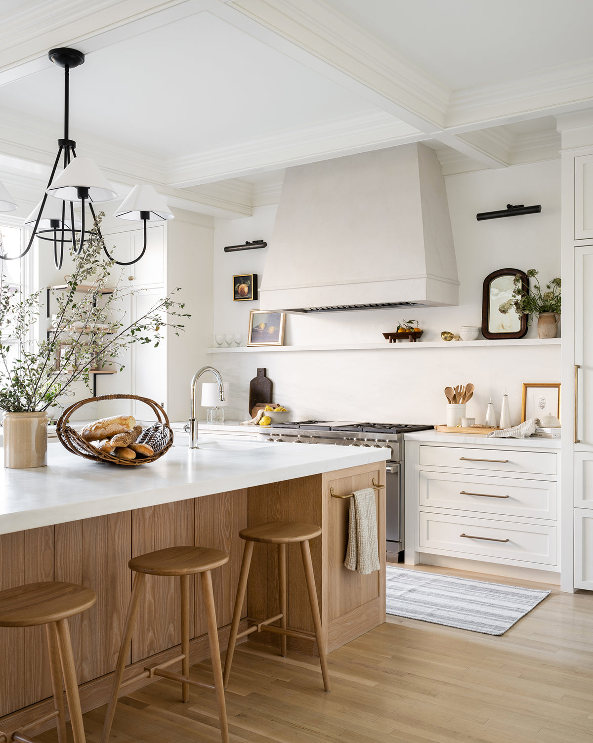 A bright, modern kitchen with light wood floors, white cabinets, a marble island topped with three Fielder Counter Stools in light oak, a bread basket, potted plants, and minimalist decor bathed in natural light by McGee & Co.