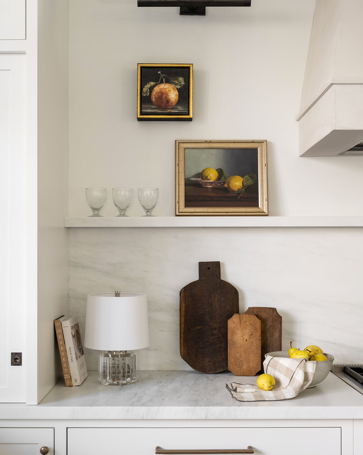 A modern kitchen with a white marble countertop, three wooden cutting boards, a bowl of lemons on a towel, the Gloomy Clementine I framed artwork evoking museum-quality still life, four glasses on a shelf, and books against the wall.