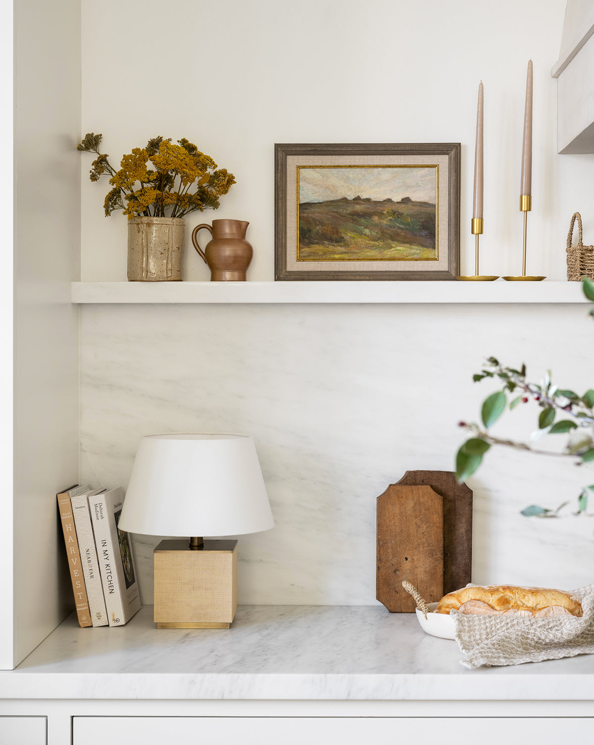 A marble shelf displays books, a square lamp, wooden decor, and a bread basket. Above it, a traditional shelf holds a vase with dried flowers, a small pitcher, two Brass Taper Holders, and a framed landscape painting.