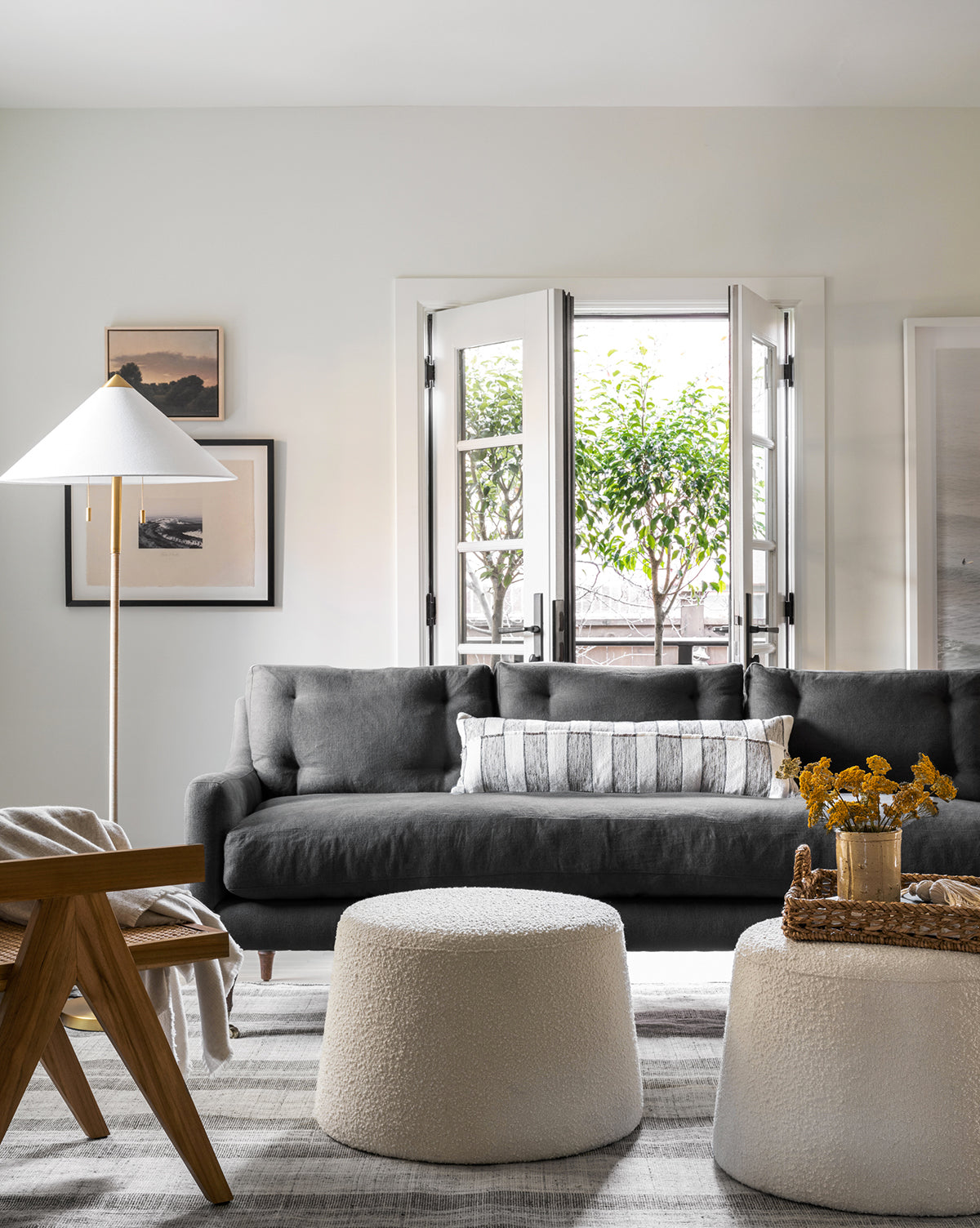 A modern living room with a Community Barden Sofa 84 featuring reeded wood legs and tufted cushions, a patterned pillow, two white textured ottomans, a wooden chair, a standing lamp, framed wall art, and French doors to trees—McGee & Co.