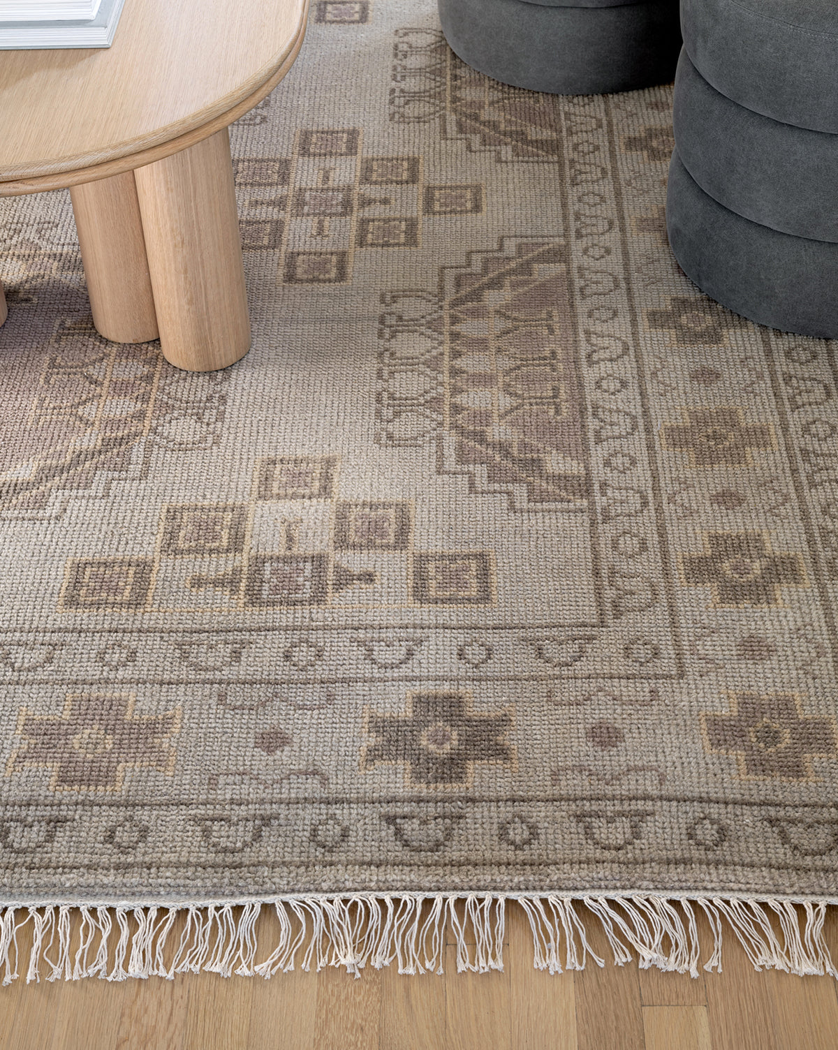 A close-up of the Kenna Hand-Knotted Wool Rug, featuring beige and brown geometric patterns with fringe, partially placed under a round wooden table beside a gray ottoman on a light wood floor, by McGee & Co.