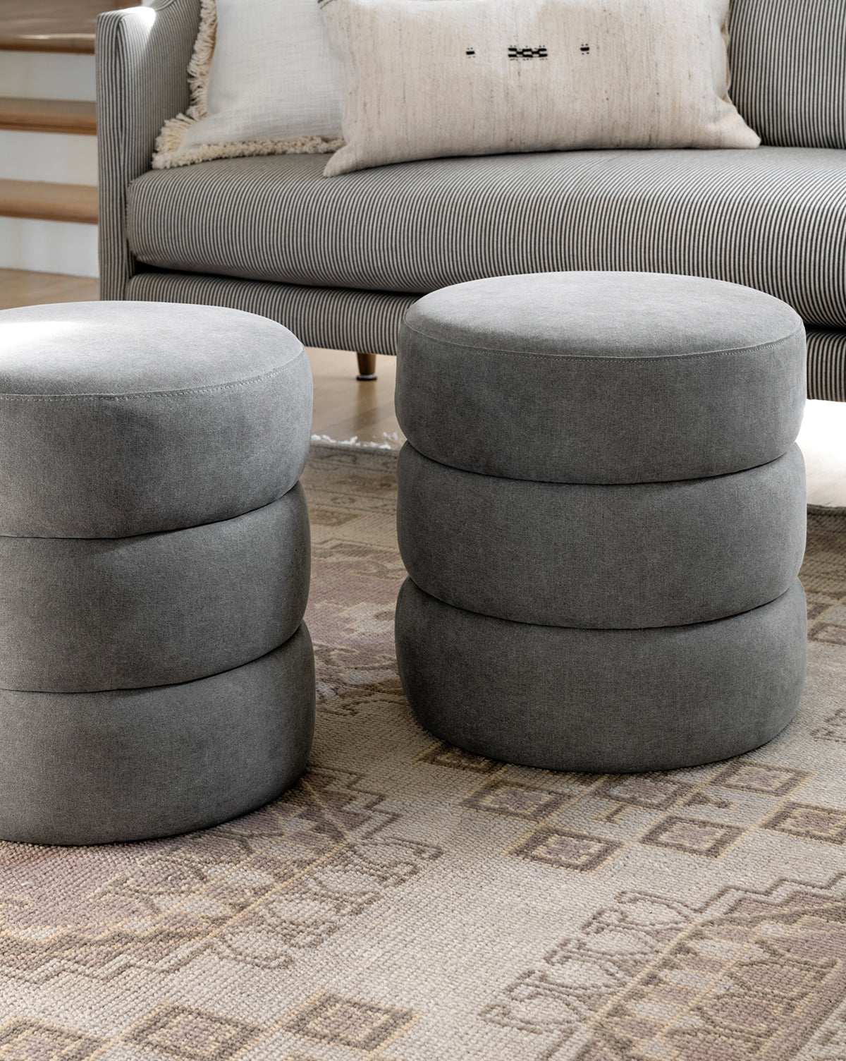Two round gray Channel Ottomans with stacked cushion-like sections sit on a patterned rug in front of a striped sofa with neutral pillows, adding midcentury flair to the space, by McGee & Co.