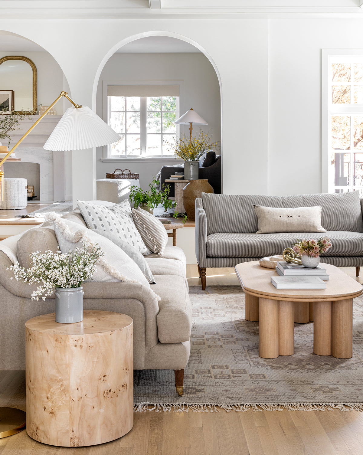 A bright, airy living room with a Makerspalm Gemma Settee 65 in high-quality fabric, patterned pillows, a round wood side table with flowers, modern coffee table, gold floor lamp, and large windows and arches for natural light — McGee & Co.