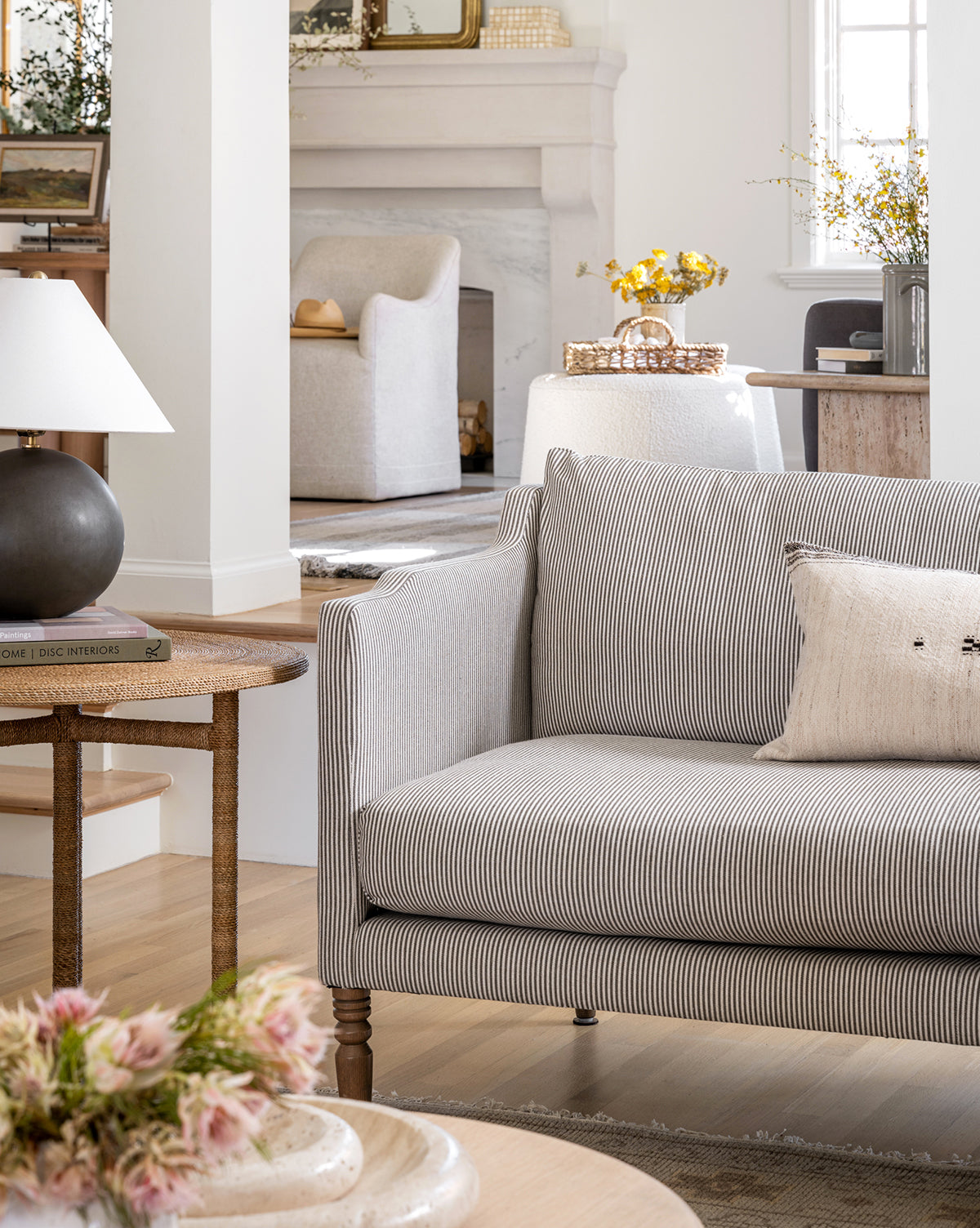 A cozy living room featuring the Makerspalm Gemma Settee 65 (Ready to Ship) in striped fabric, a beige pillow, side table with lamp and books, fresh flowers, fireplace, and another chair bathed in soft natural light. McGee & Co.