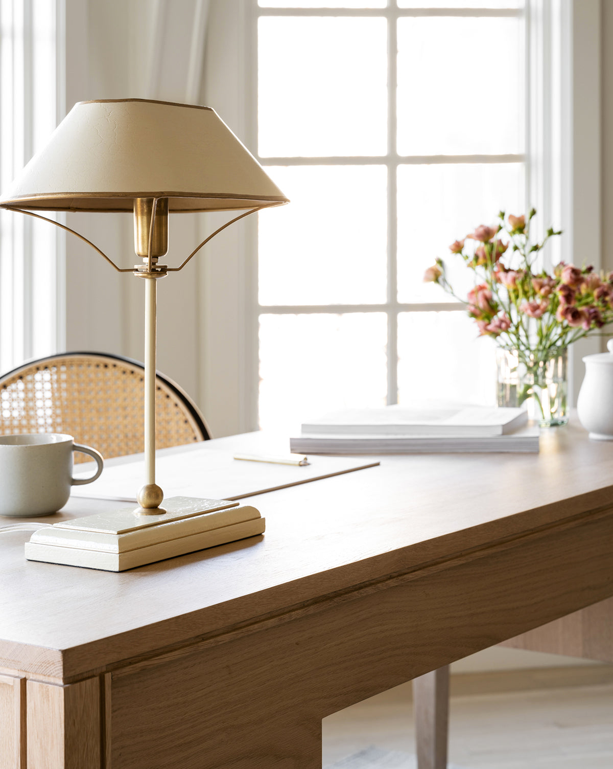 A bright, minimalist desk with the elegant Daphne Table Lamp, a white mug, stacked books, and a vase of pink flowers. Natural light flows through large windows, creating a calm and inviting workspace atmosphere by McGee & Co.