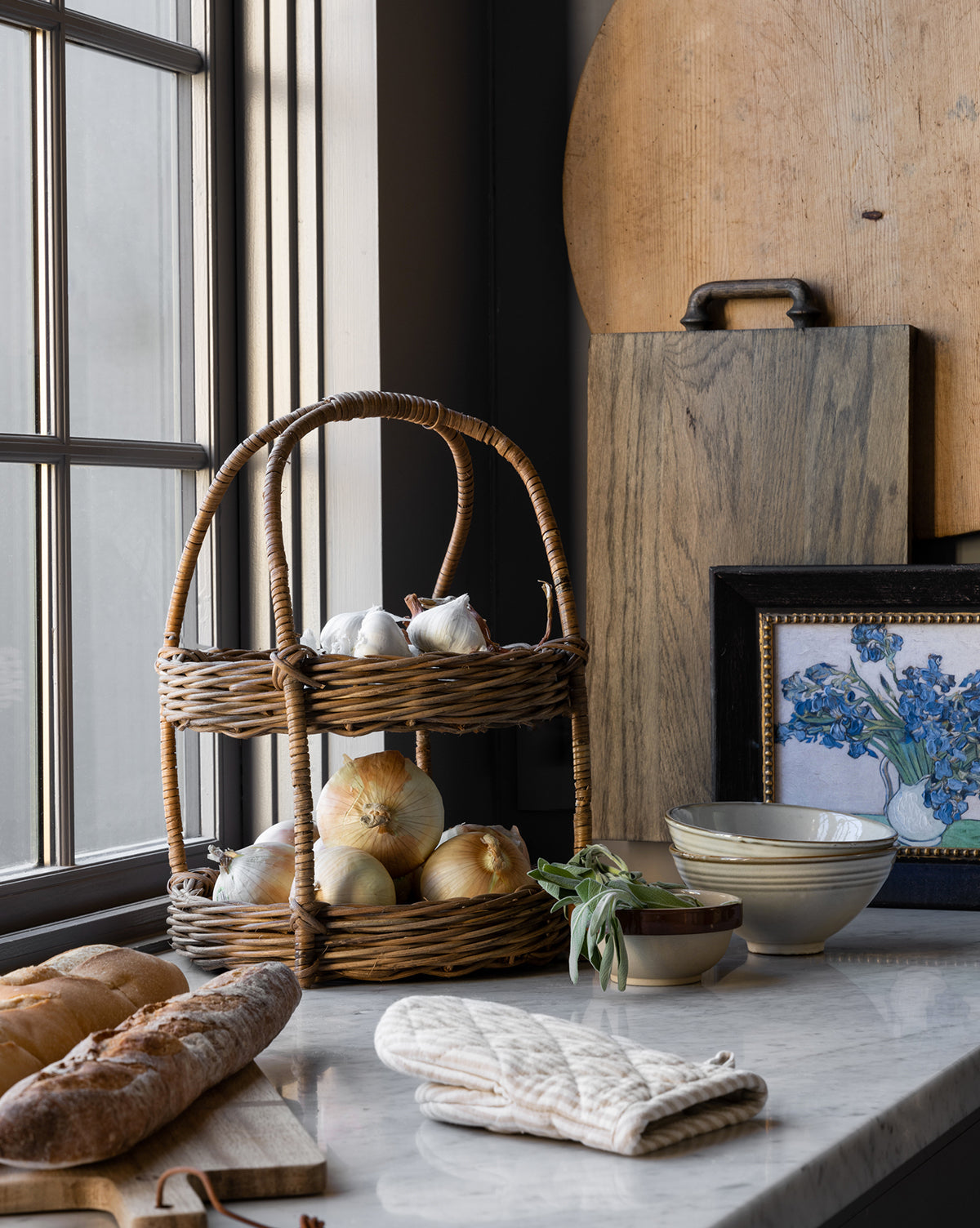 A kitchen countertop displays a two-tiered wicker basket with onions and garlic, a loaf of bread, the Fordham Board (solid oak), ceramic bowls, floral artwork in a frame, and a white oven mitt by the window, by McGee & Co.