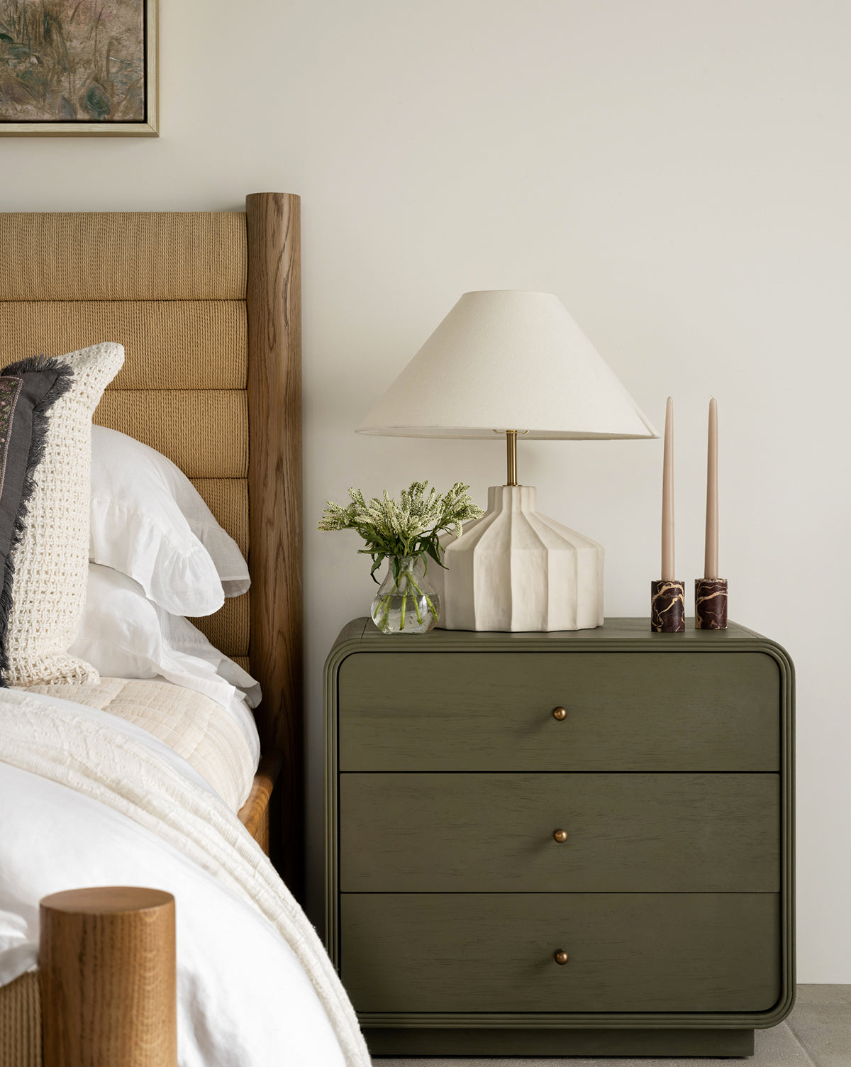 A neatly made bed sits beside a green three-drawer nightstand, which is topped with a Veneto Table Lamp, two tall candles in holders, and a small vase of flowers, all set against a plain white wall by McGee & Co.
