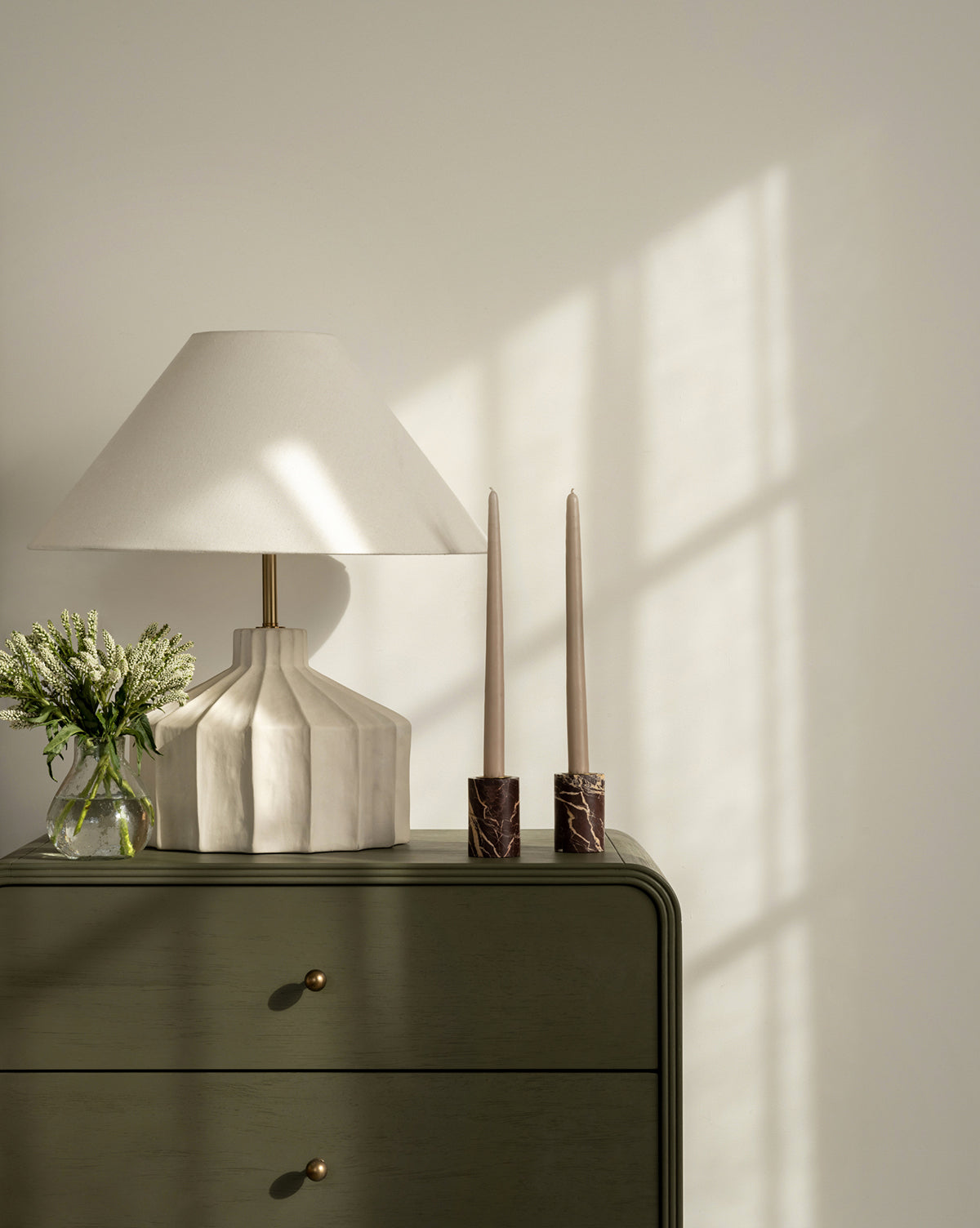 A soft-lit scene features a white table lamp, a small vase of flowers, and Perrin Marble Taper Holders (Set of 2) with tall candles on a green dresser, windowpane shadows cast on the light-colored wall behind.