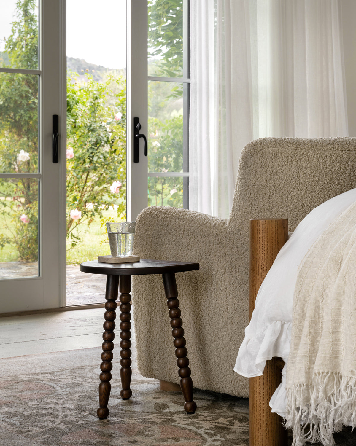 A cozy beige armchair and a white bed sit by glass doors, complemented by the Alice Wood Stool in ash. Sheer curtains and natural light create a serene, inviting atmosphere with views of the lush garden by McGee & Co.
