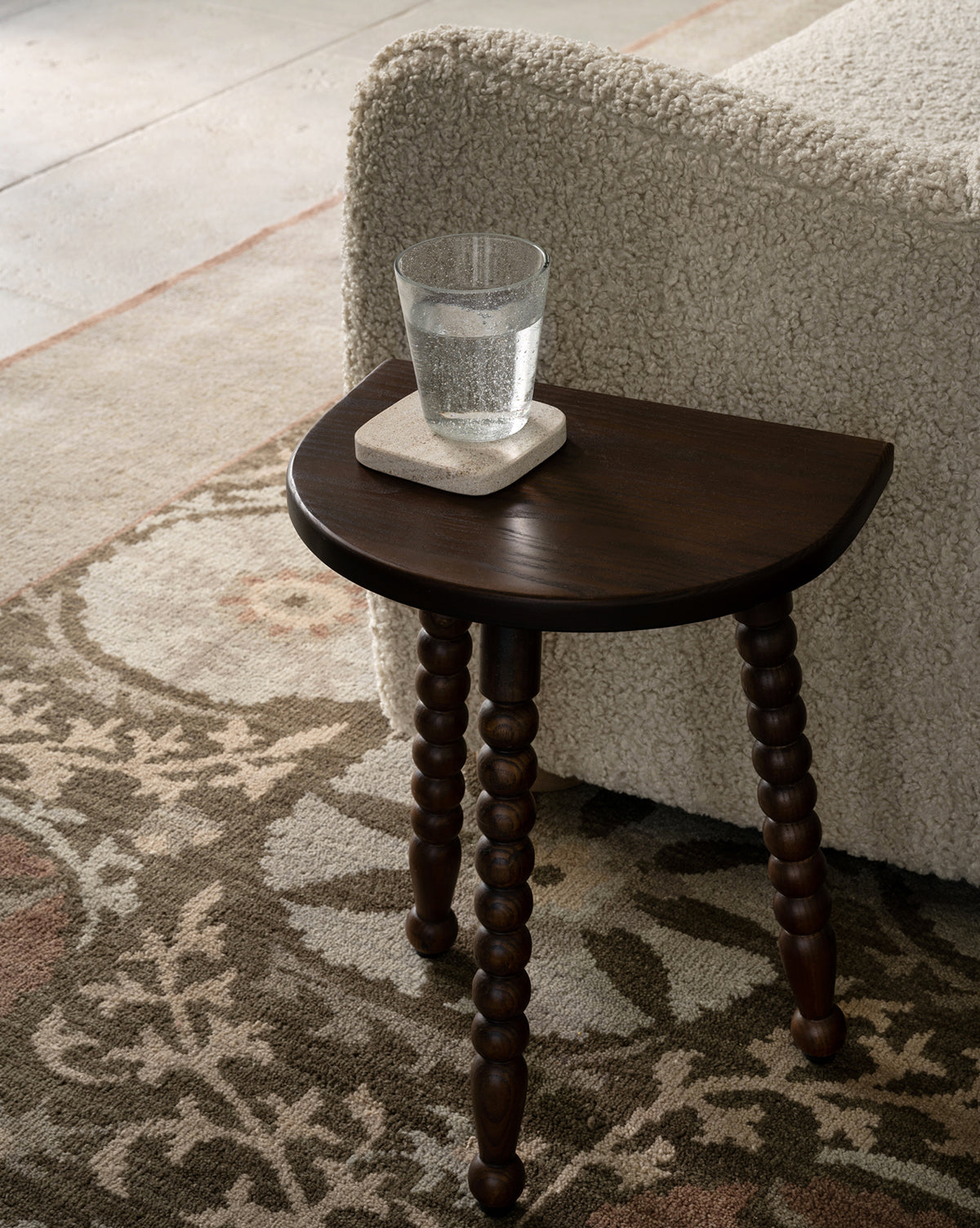 A small dark wood side table with carved legs holds a glass of water on a stone coaster beside a textured cream sofa, all set atop the Suzani Hand-Knotted Wool Rug, by McGee & Co.
