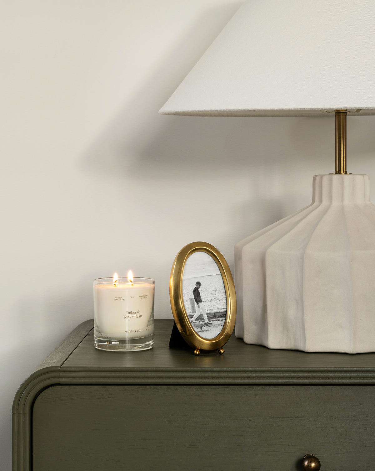 A bedside table with a lit candle, a Blanche Picture Frame displaying an oval black and white photo, and a modern lamp featuring a white shade and geometric base.