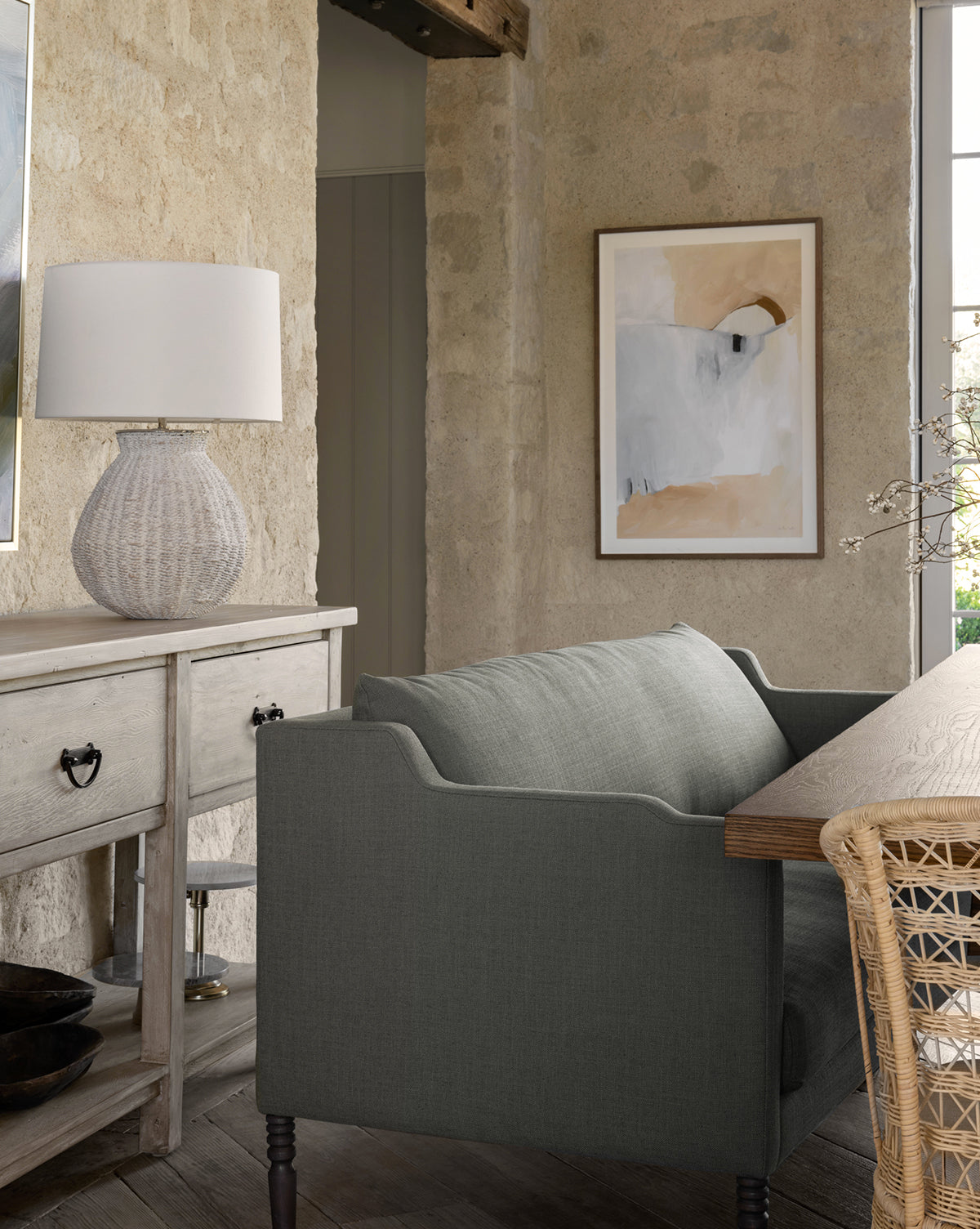 A cozy living room features the Makerspalm Gemma Settee 65 in high-quality fabric, paired with a light wooden sideboard, textured white lamp, abstract wall art, and a rustic stone wall for a warm, inviting atmosphere—finished with thoughtful touches inspired by McGee & Co.