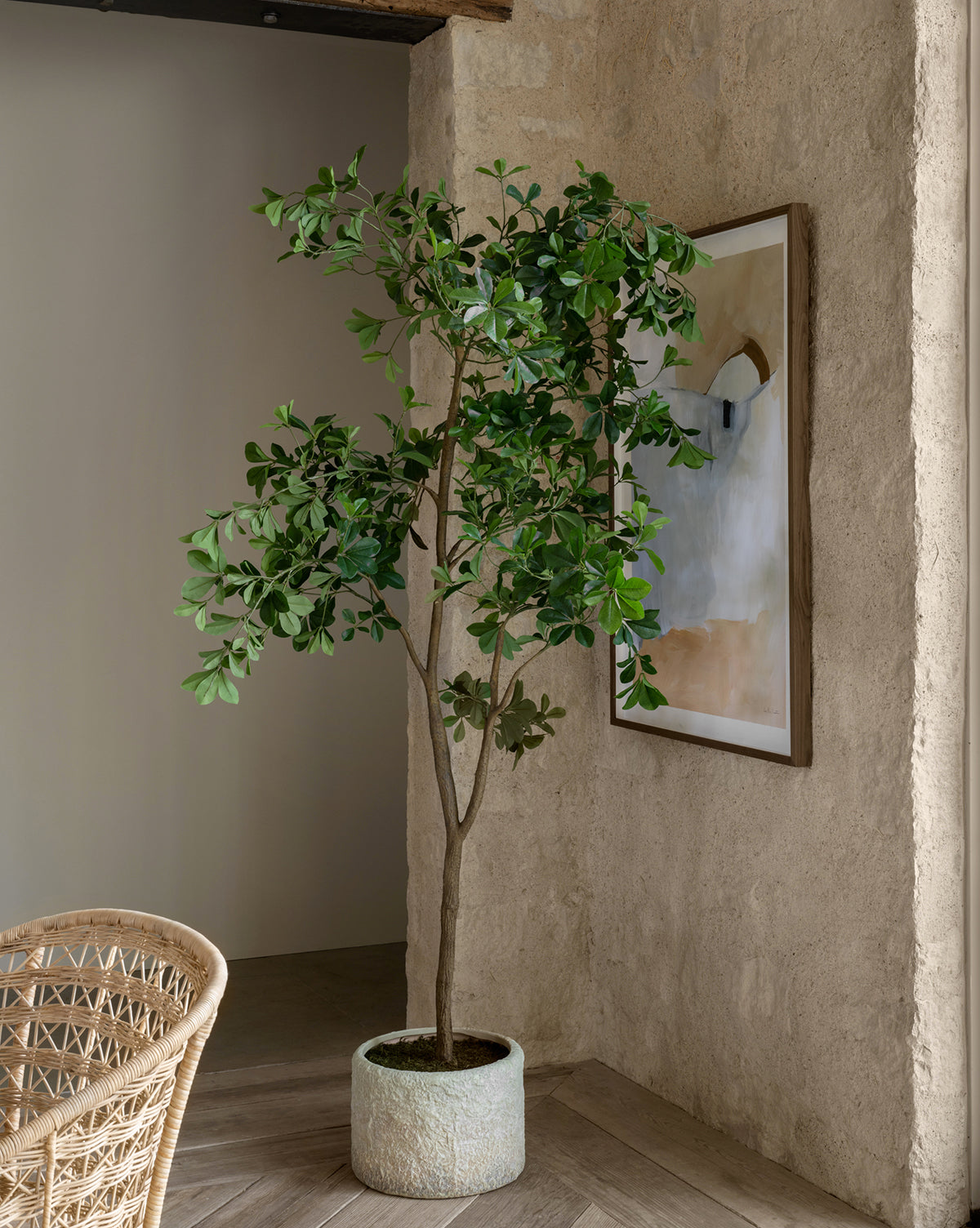 A tall Shady Lady Faux Tree with vibrant green leaves is placed in the corner of a rustic room with textured beige walls, beside a woven chair and a framed abstract painting.