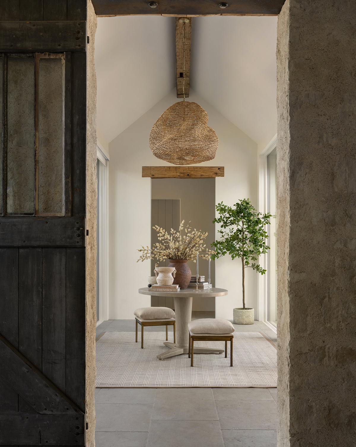 A rustic dining area with a round table, two stools, a large vase with branches, a potted tree, and organic lighting from the Andorra Wicker Pendant. The room features stone walls, wooden beams, and neutral tones, by McGee & Co.