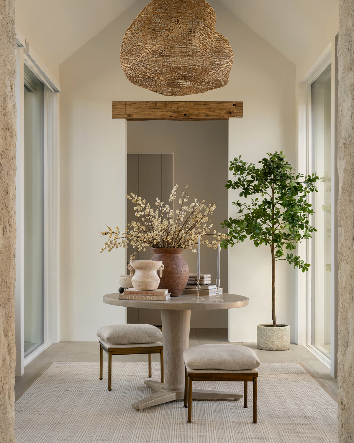 A bright room with beige walls includes a small round table with vases and books, two cushioned stools, and the Shady Lady Faux Tree in a ceramic pot. Natural textures and a woven pendant light create an airy, calming atmosphere.