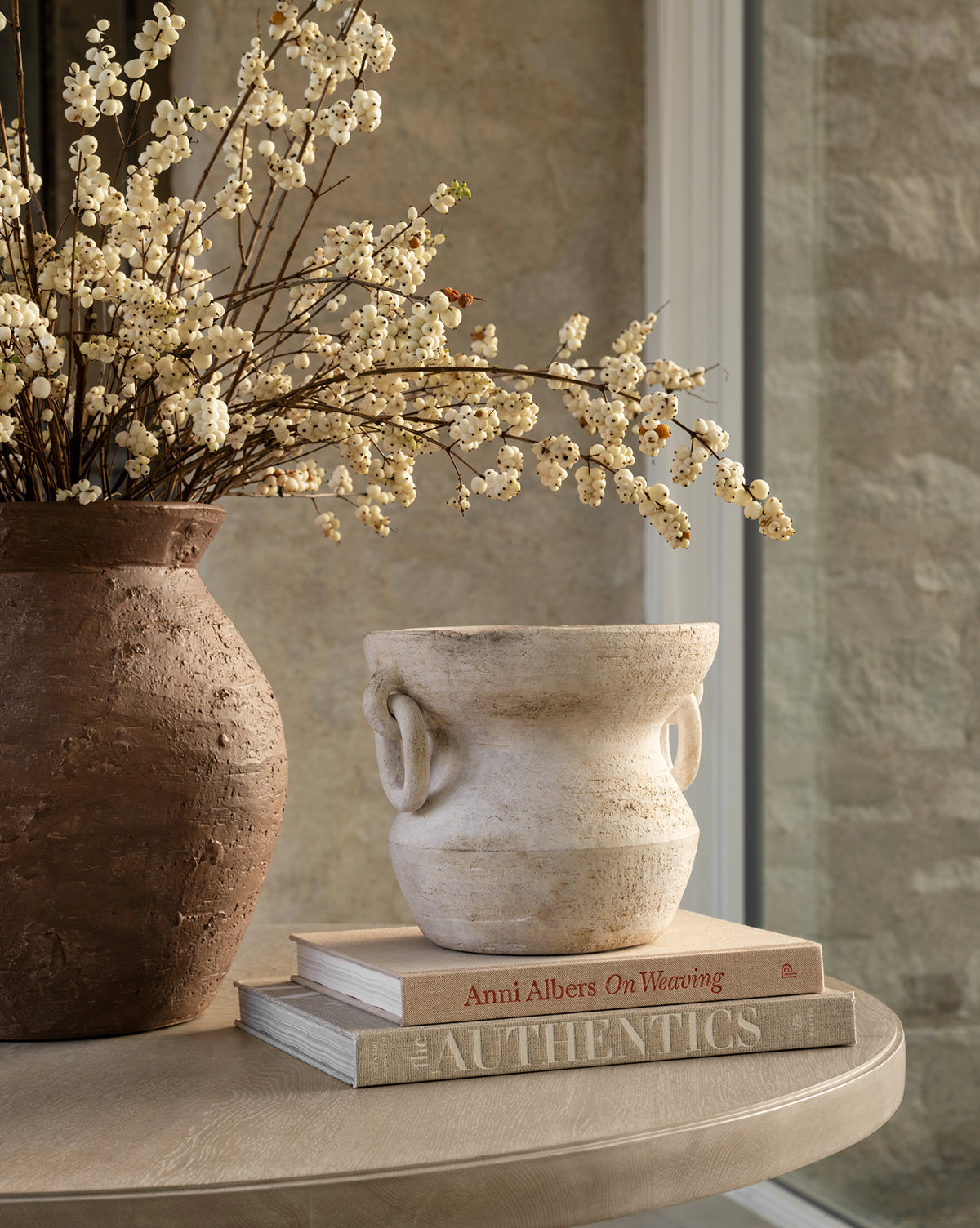 A rustic round table holds the Arnette Terracotta Vase with white flowers, a textured white pot, and two stacked books—“Anni Albers on Weaving” and “The Authentics”—beside a stone wall and window.