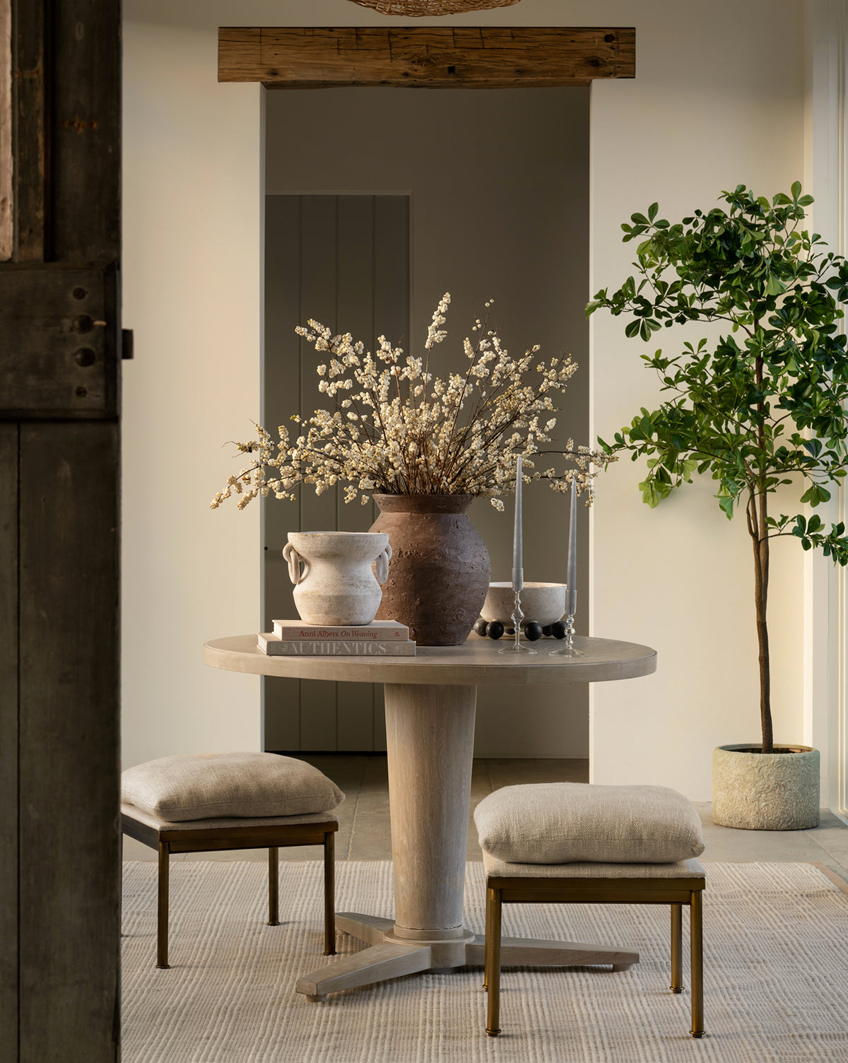 A round wooden table with two cushioned stools holds a Demetria Terracotta Vase of white flowers, a ceramic pitcher, books, and two tall candles. A potted tree stands by the wall in a softly lit, minimalist room.