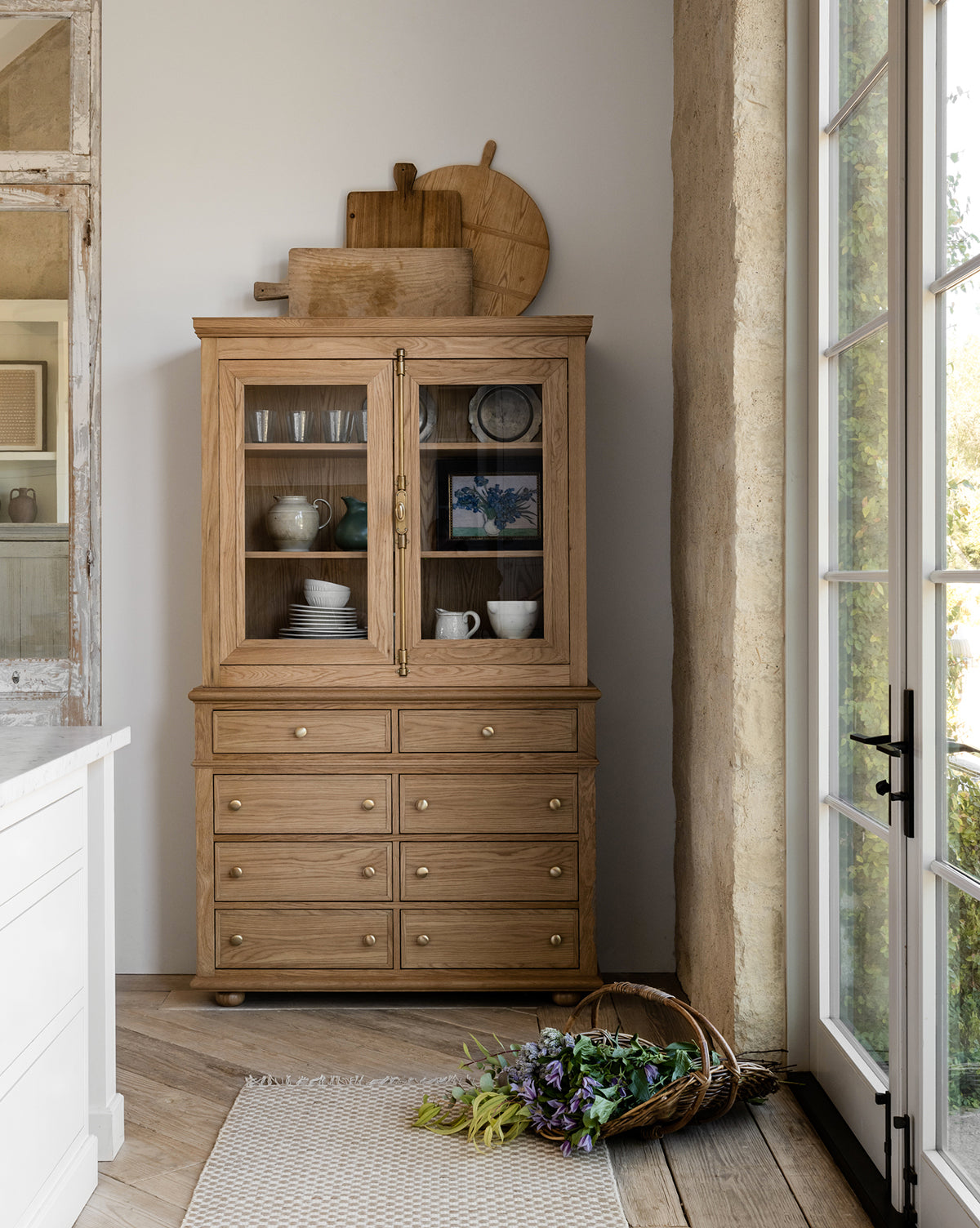 The Heath Oak Cabinet, featuring drawers and glass doors, showcases dishes and decor. Nearby, a basket of fresh flowers sits on a woven rug as sunlight pours through large glass doors beside this elegant traditional piece by McGee & Co.