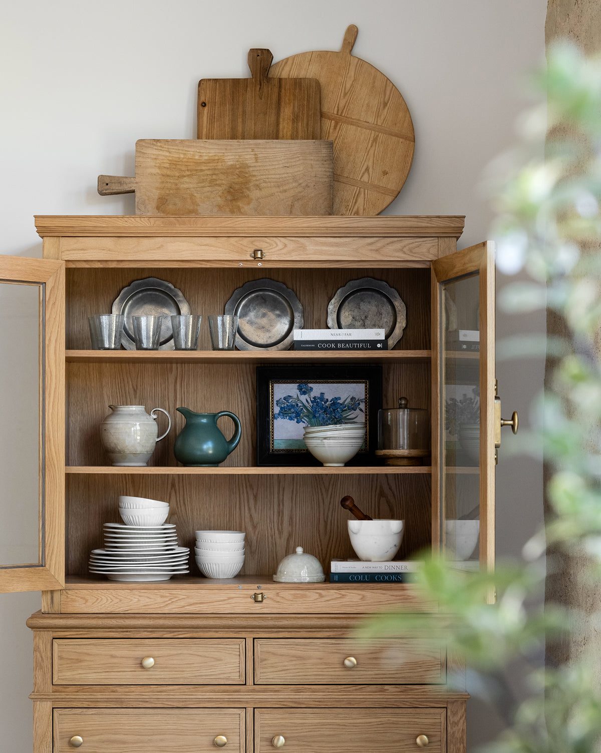 A wooden cabinet with glass doors displays plates, bowls, cups, a green teapot, books, and glassware. On top, cutting boards sit beside a black and gold wood frame featuring the artwork Irises by Van Gogh. A plant is blurred in front.