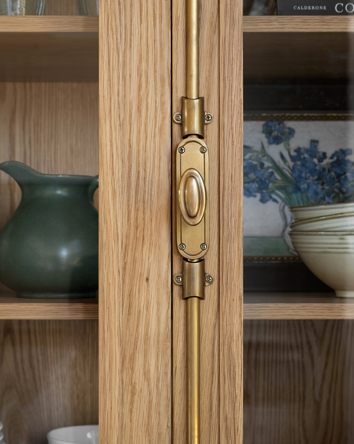 Close-up of a brass handle on the Heath Oak Cabinet, featuring shelves inside that display a green pitcher, striped bowl, white dishes, and a framed floral painting by McGee & Co.
