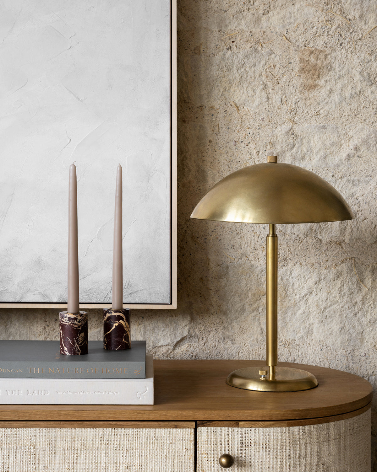 A modern table setup features beige candles in Perrin Marble Taper Holders (Set of 2), stacked books, and a lamp with a brass cap on a wooden cabinet, set against a textured stone wall and a large minimalist painting.