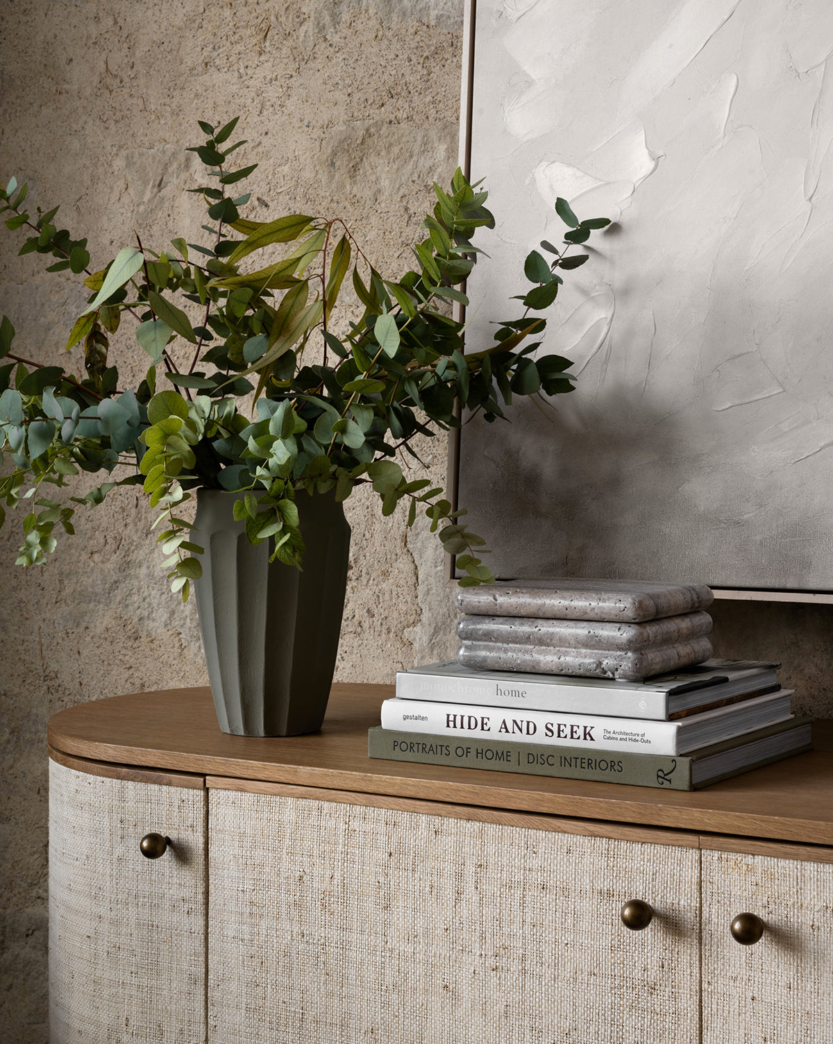 The Frida Vase, hand-glazed and filled with leafy branches, sits on a wooden cabinet beside stacked books and folded textiles, set against a textured wall with abstract artwork in the background.