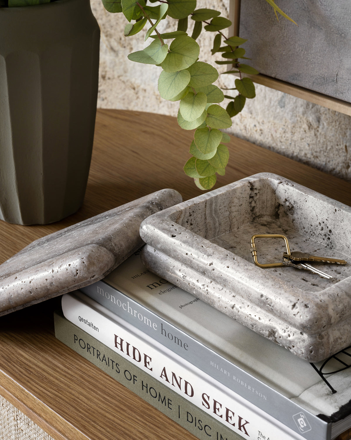 The Ives Stone Box, with its lid set aside, holds a key and ring. This decorative square marble box rests on wood next to stacked books and a green vase filled with eucalyptus leaves.
