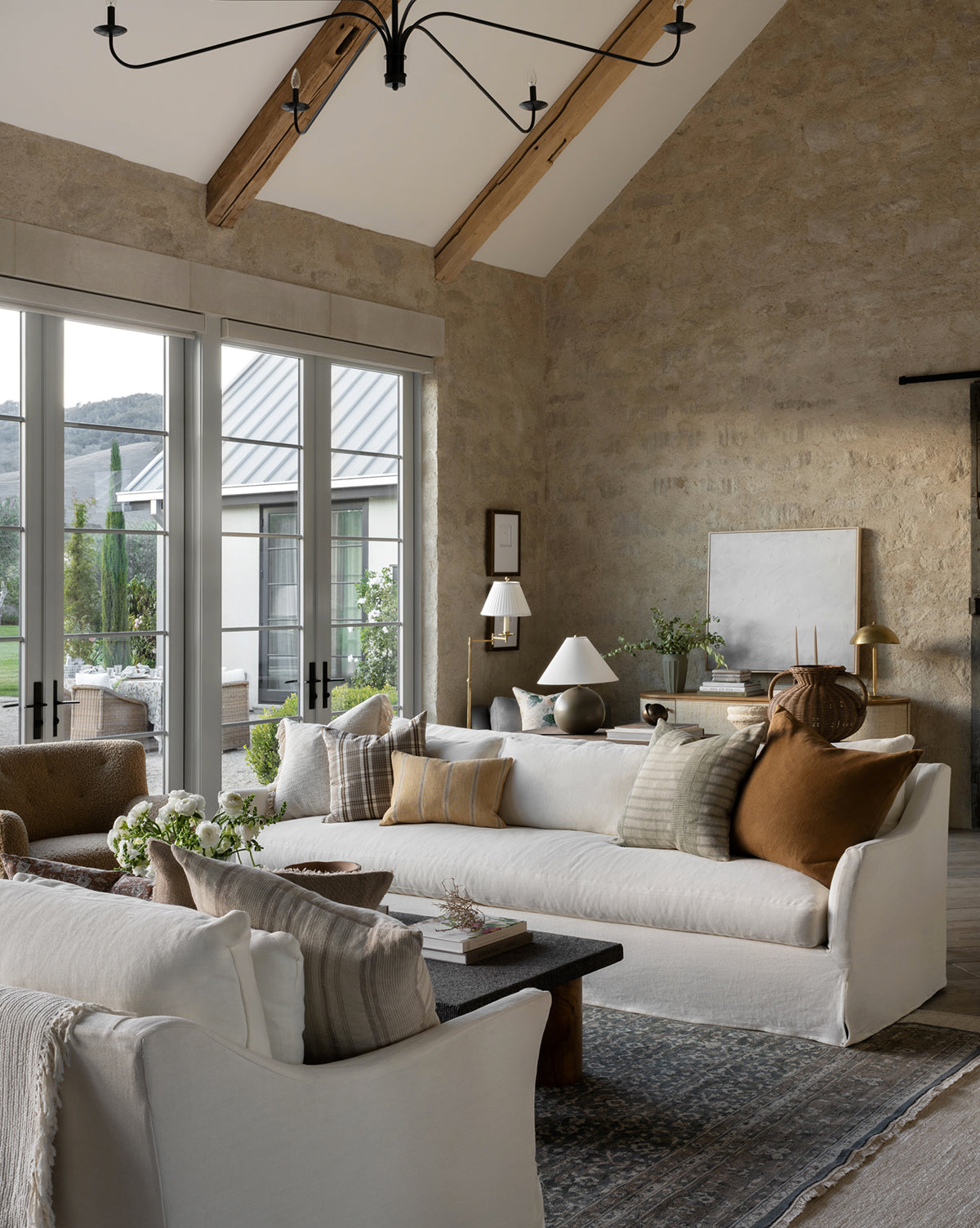 A modern living room features high vaulted ceilings, beige stone walls, large windows overlooking greenery, white sofas with neutral and brown pillows, a black coffee table, and the Marta Hand-Knotted Wool Rug accenting the space, by McGee & Co.