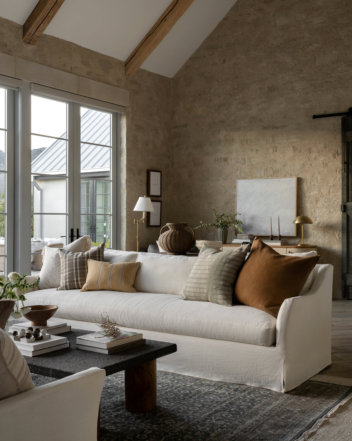 A cozy living room features the Rowe Fine Furniture Wilhelmina Classic Slipcover Sofa (85-110) with neutral and brown pillows, a dark coffee table, greenery, large windows, stone walls, wood beams, minimalist decor, and abundant natural light, blended seamlessly with pieces from McGee & Co.