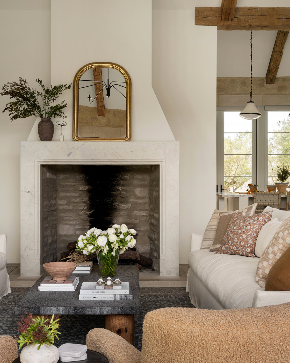 A cozy living room in neutral tones features a stone fireplace with the Giada Wall Mirror above it, a dark coffee table topped with flowers and books, two sofas with patterned pillows, and exposed wooden ceiling beams for an antique touch, by McGee & Co.