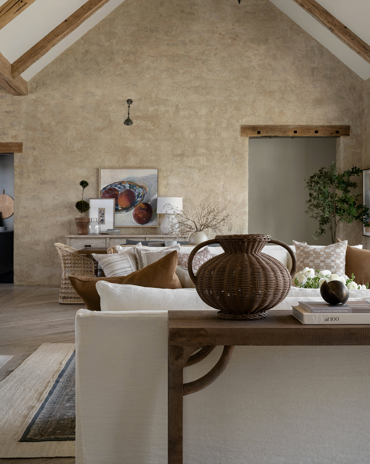 A cozy, neutral-toned living room with rustic textured walls, exposed wooden beams, a white sofa, brown armchairs, the Antonia Woven Vase, artwork on the back wall, and potted plants adding warmth and greenery.