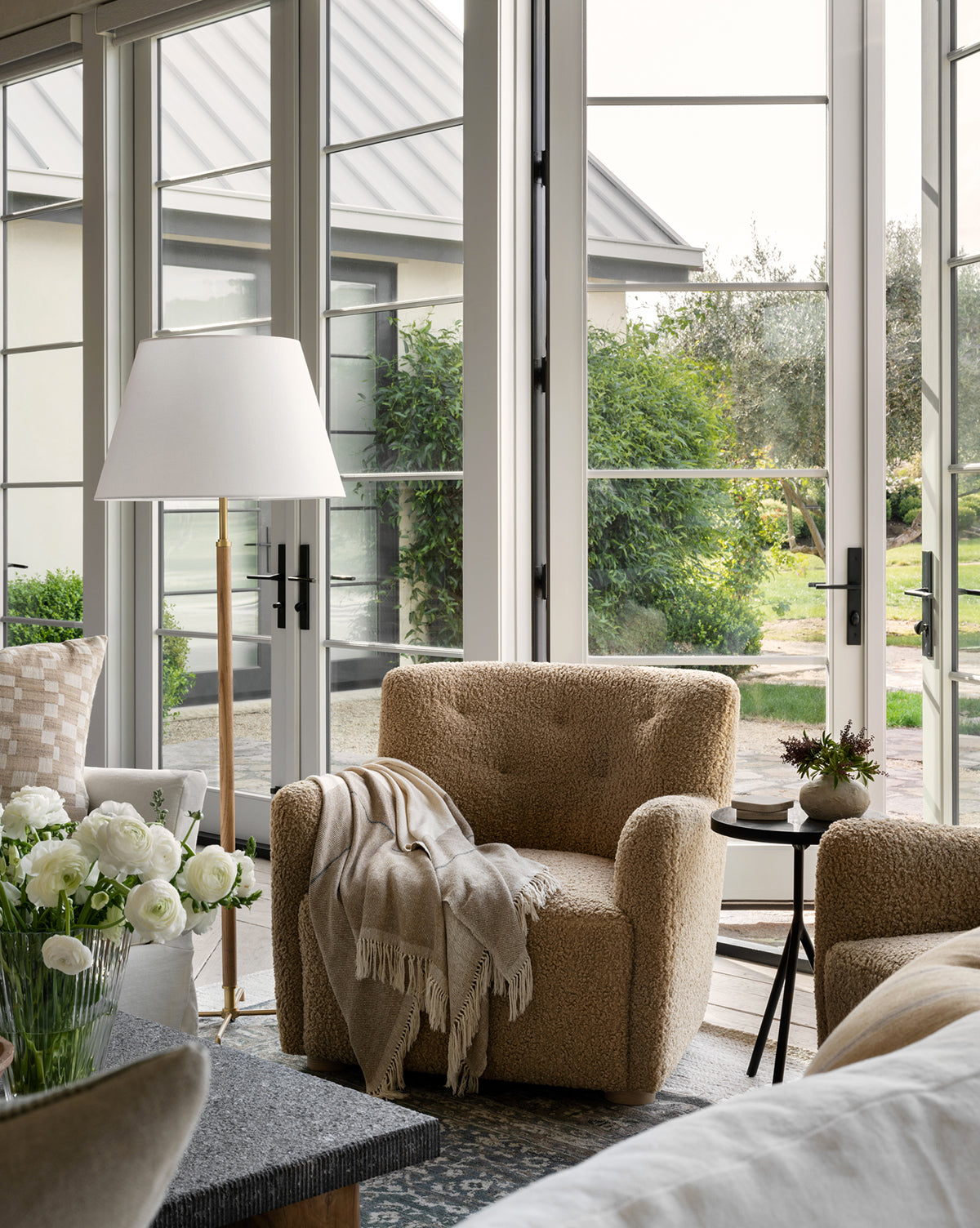 The Magda Lounge Chair (Ready to Ship) in beige, draped with a light throw, sits by a round side table and floor lamp in a bright living room near glass doors opening to a green garden. White flowers adorn a nearby table, by McGee & Co.